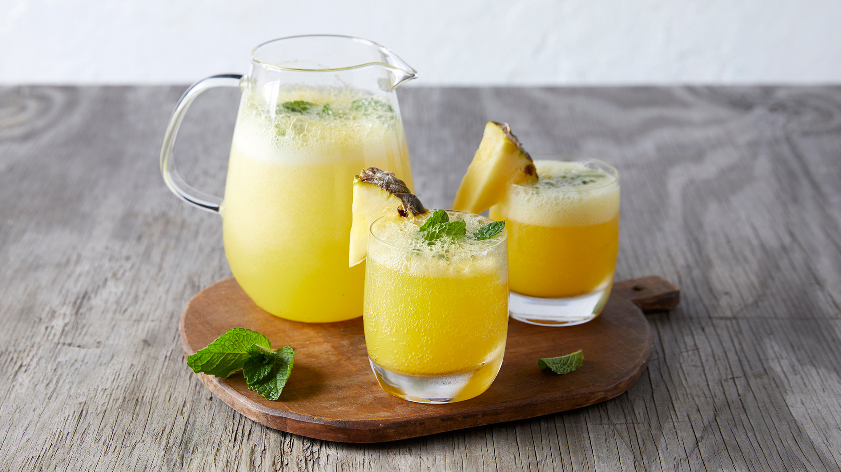 A jug and two glasses of golden Passion & Pineapple Crush sit on a wooden board, decorated with wedges of pineapple and mint.