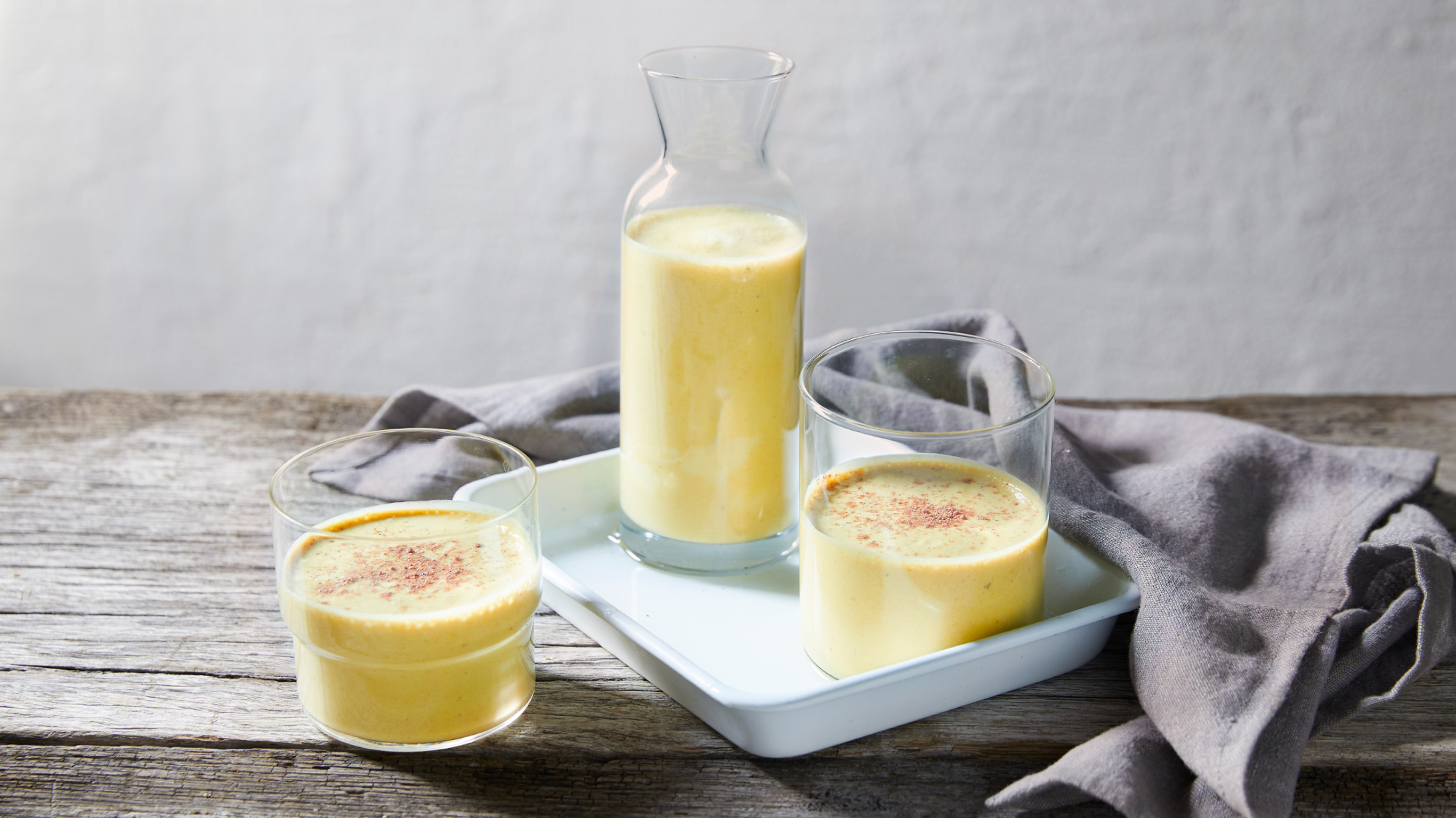 A glass and pitcher of Centr's Creamy Coconut & Turmeric Smoothie sit on a white tray, with a second glass of smoothie on the side.