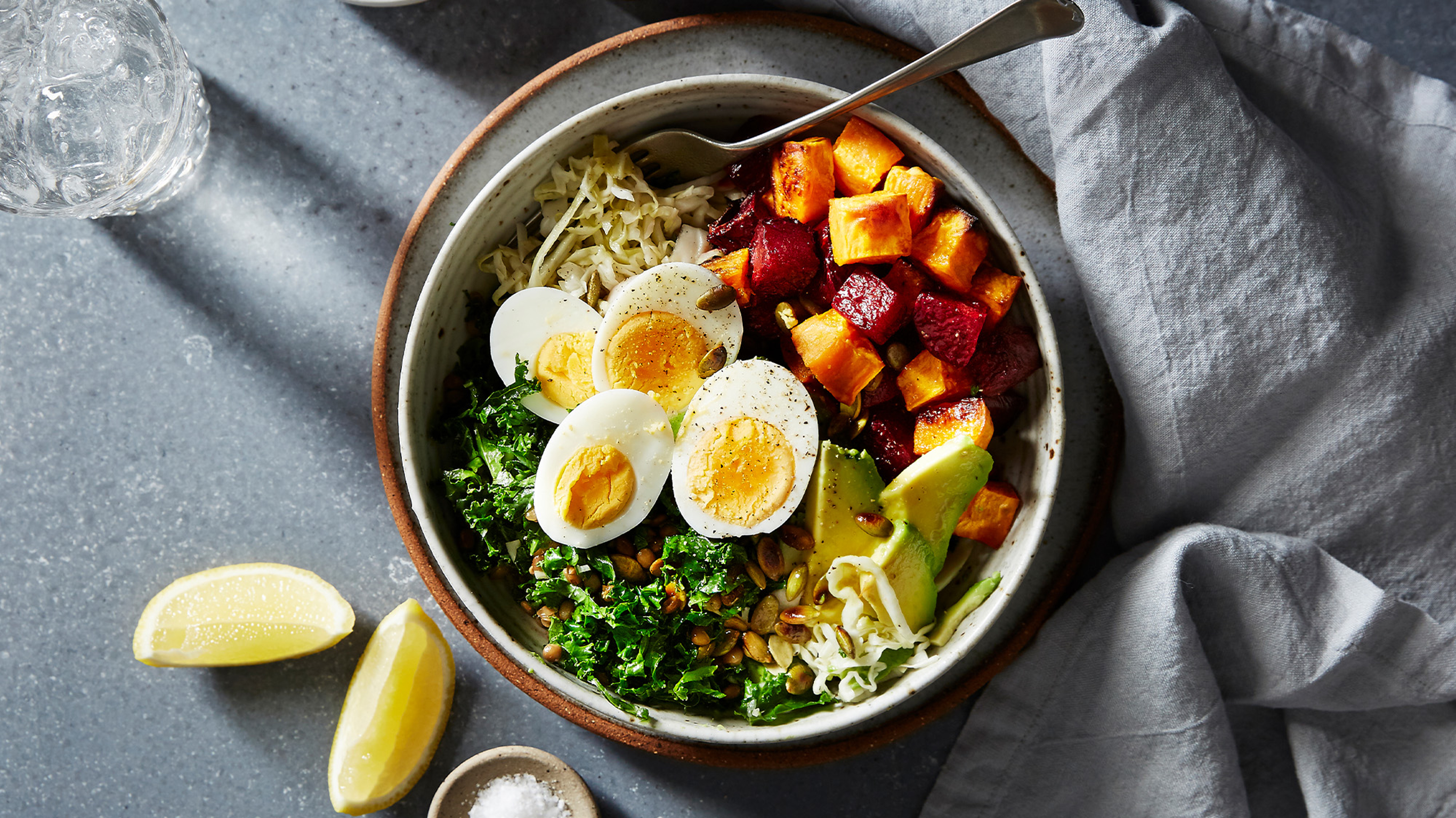 A bowl of Centr's Roast Veggie Protein Bowl with Eggs rests on a stone bench with lemon wedges and a small bowl of rock salt on the side.