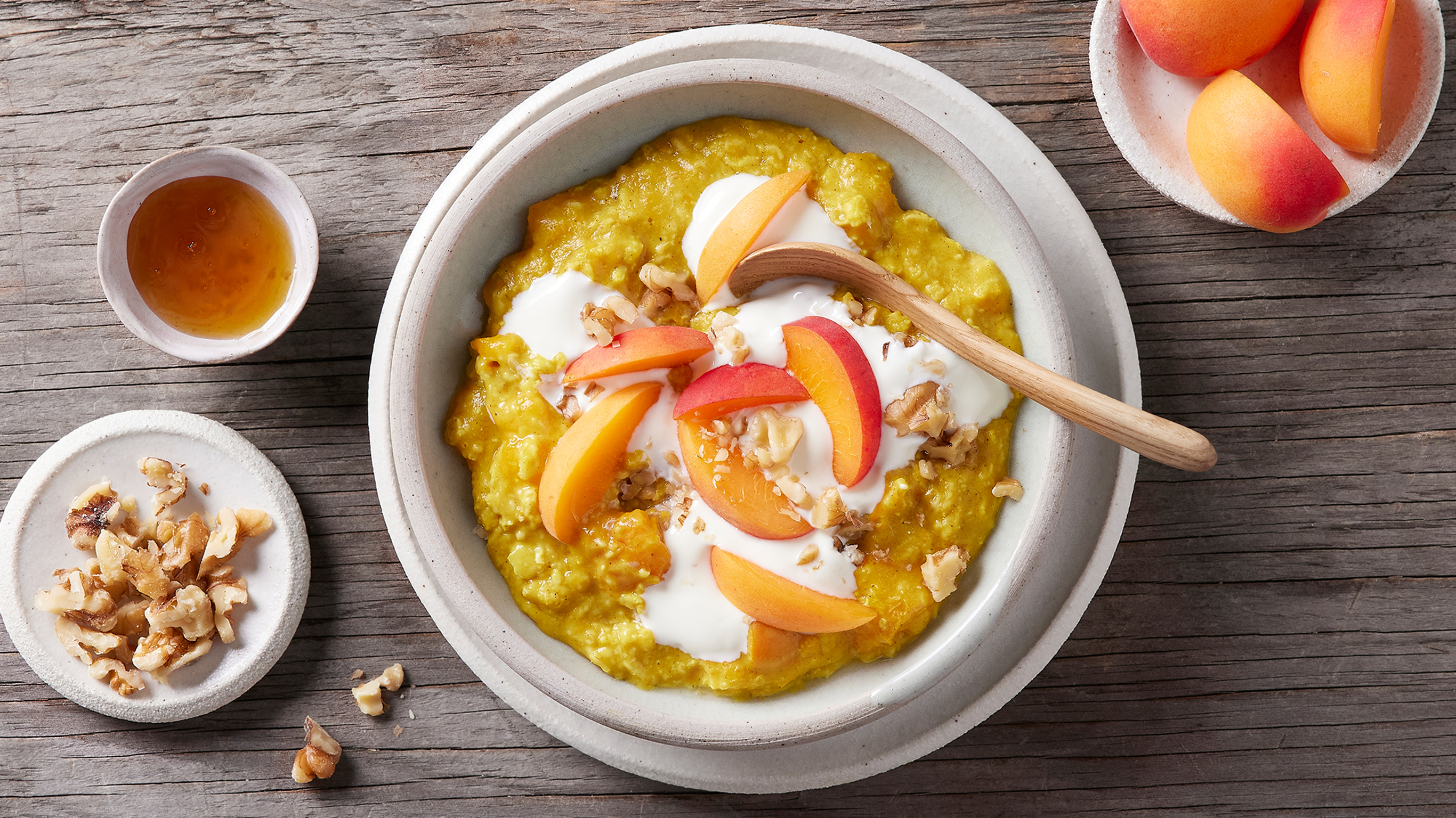 A bowl of Centr's Golden Oatmeal, topped with yogurt and sliced apricots, sits on a wooden table, with a wooden spoon resting in the bowl.