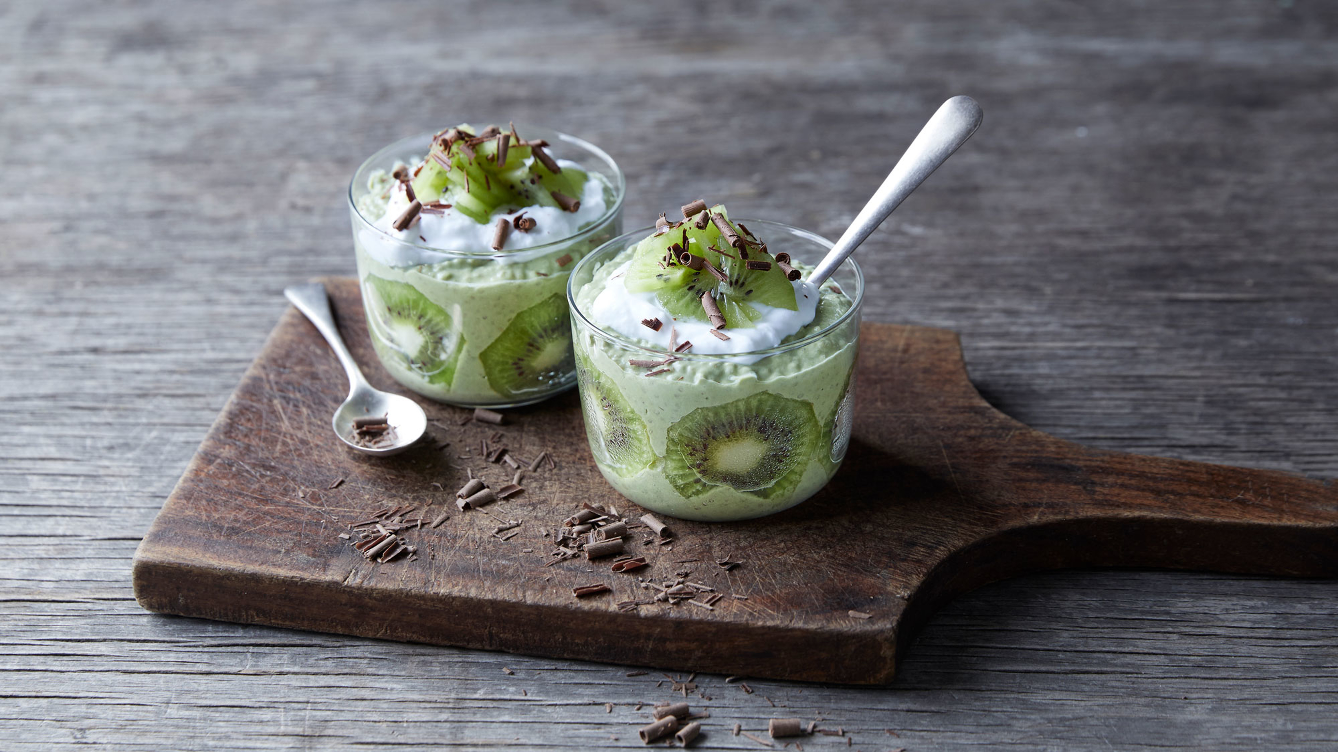2 small glass bowls of Centr's Kiwi Matcha Pudding sit on a wooden serving board with dark chocolate flakes sprinkled around.