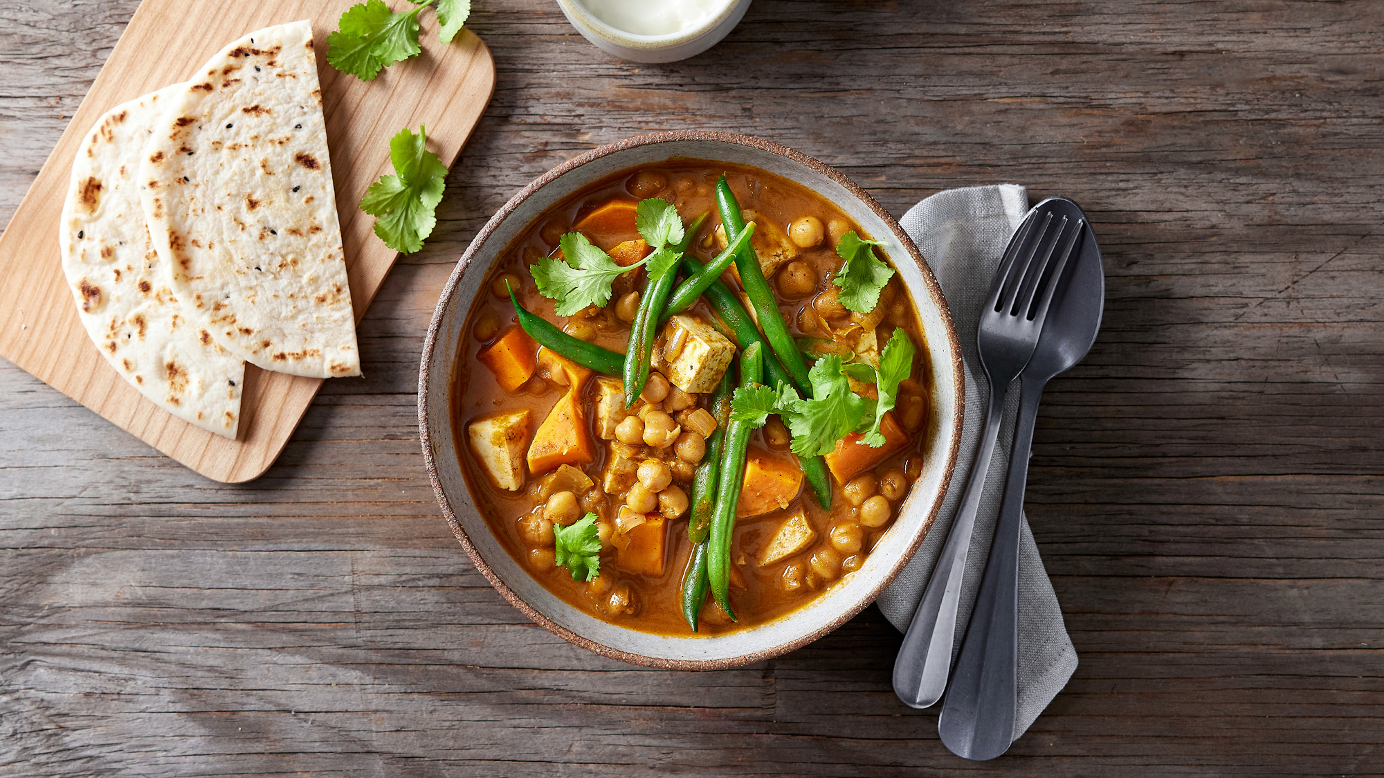 A bowl of Centr's Creamy Vwggie Korma Curry rests on wooden table with a side serve of flatbreads.