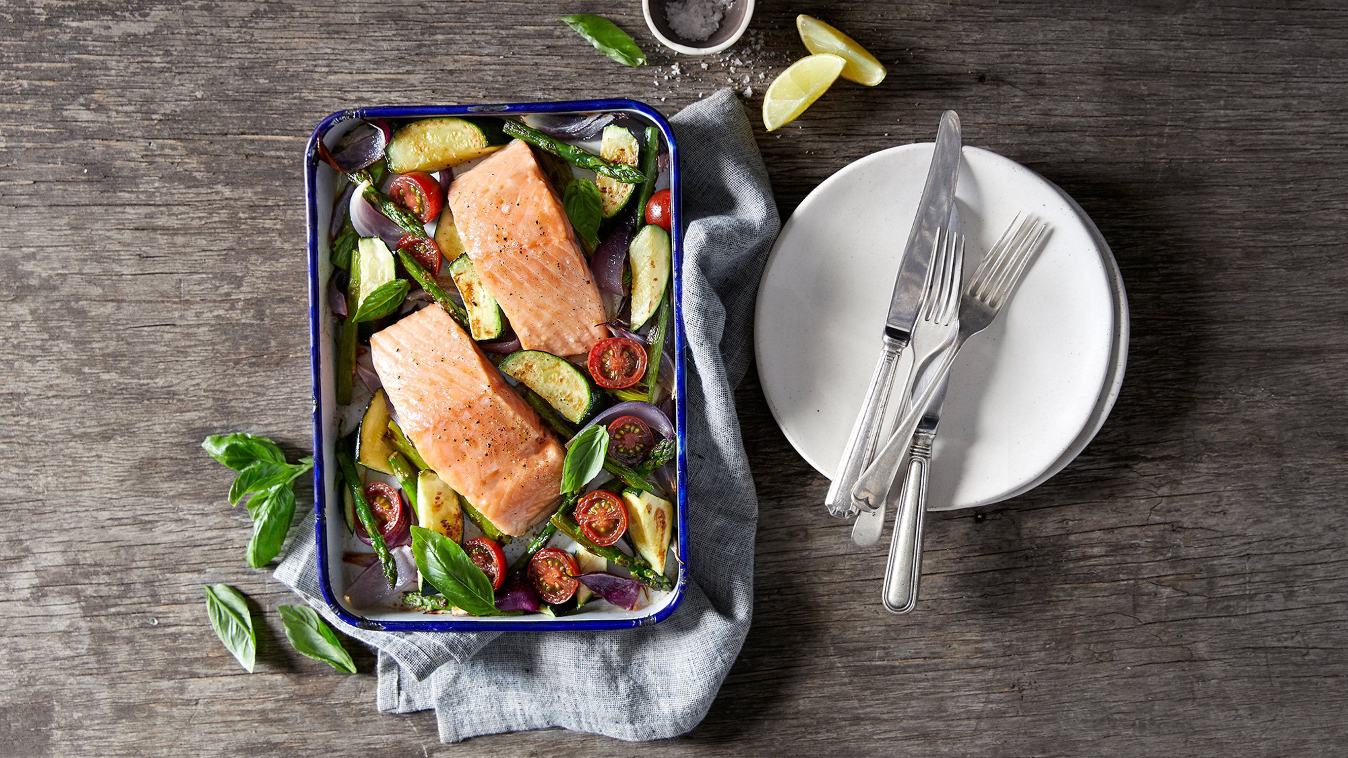 A tray of Centr's Sheet-pan Salmon & Roasted Vegetables sits on a grey linen napkin on a wooden table, ready for serving.