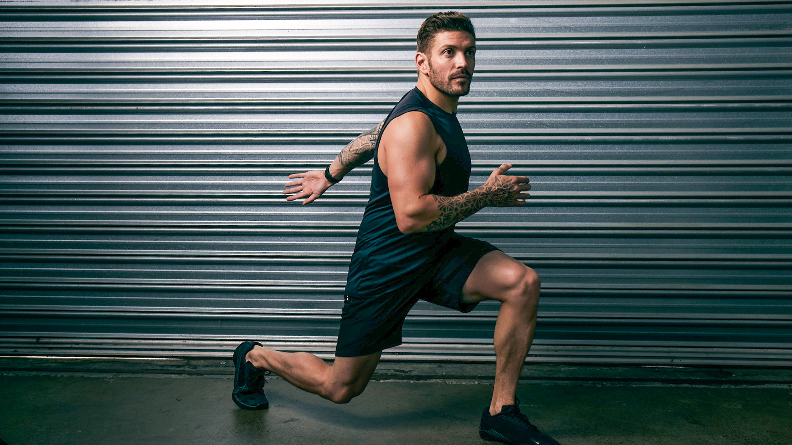 Centr trainer Luke Zocchi, wearing black gym gear, performs a lunge in front of a closed, grey roller door.