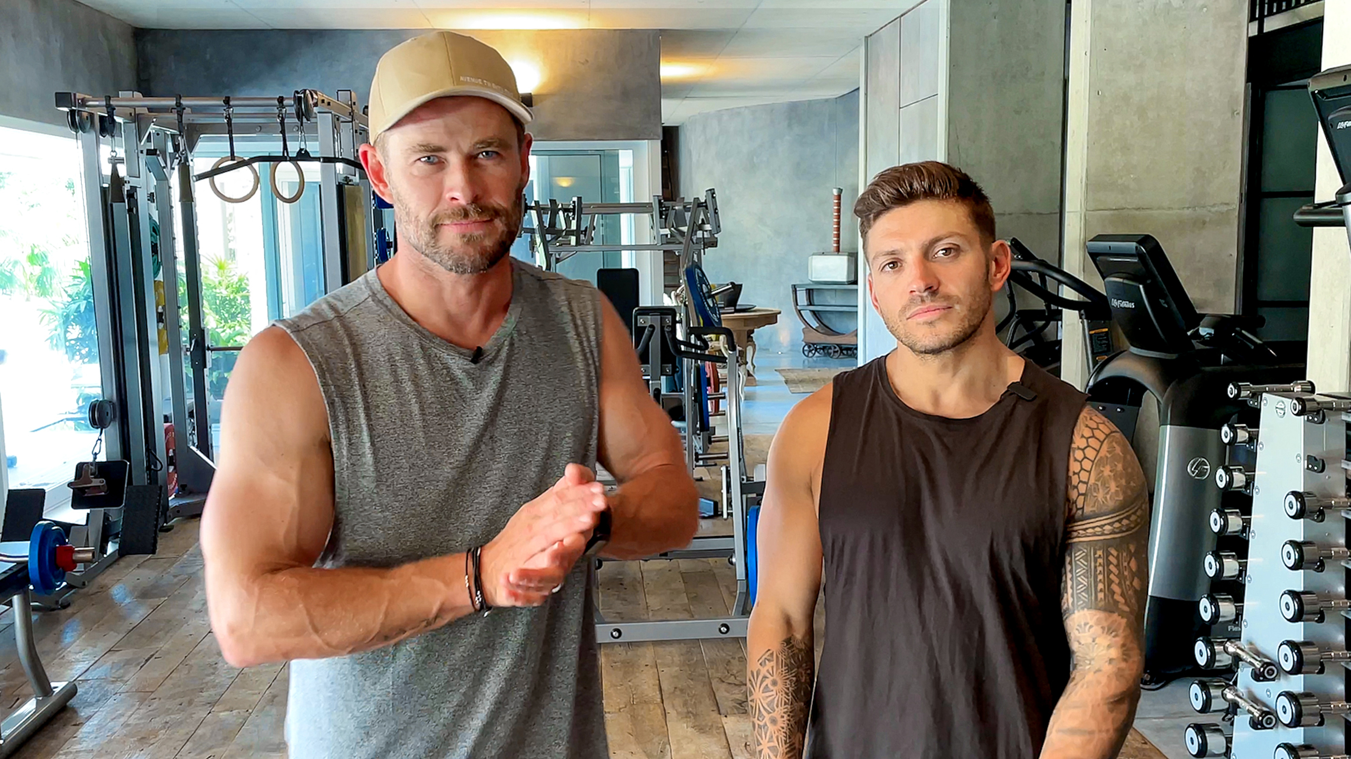 Chris Hemsworth and Luke Zocchi stand in a home gym, looking at the camera, ready to workout.