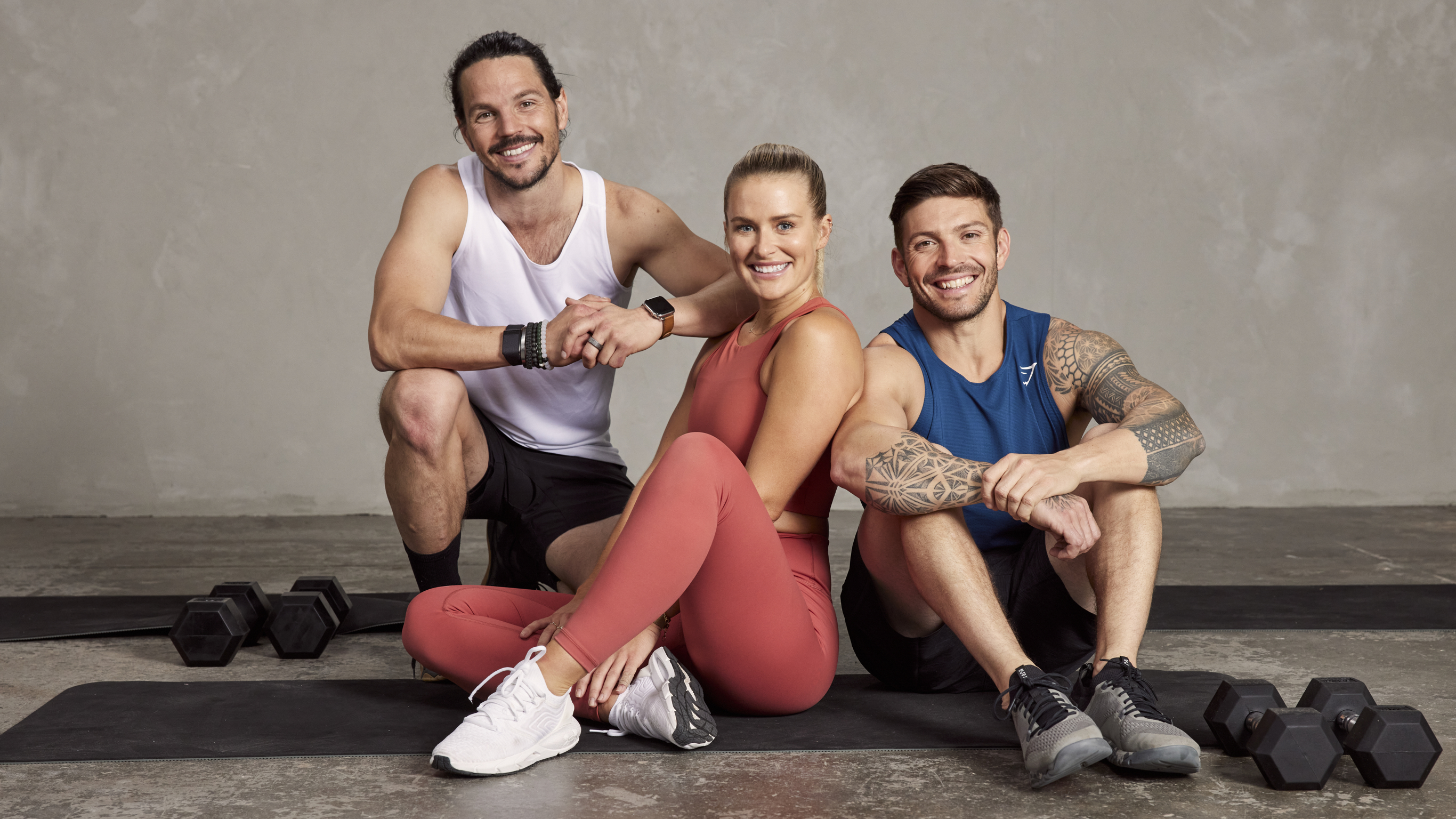 Centr trainers Dan Churchill, Alexz Parvi and Luke Zocchi sit for a group photo, on the gym floor, with gym mats and dumbbells nearby.