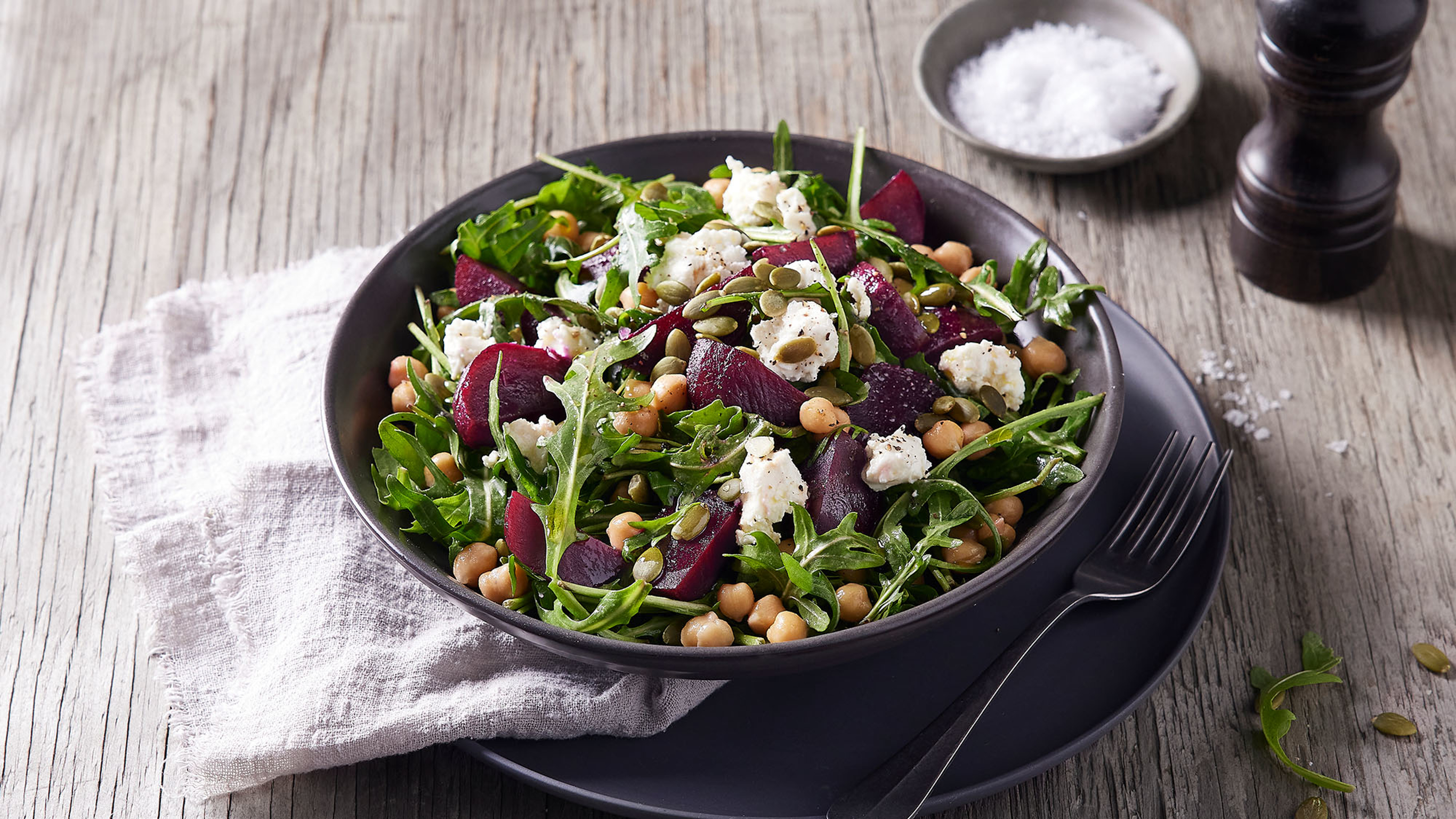 A charcoal grey coloured bowl of chickpea & beet salad with goat's cheese rests on a matching plate, with a light grey linen napkin and fork on the side.