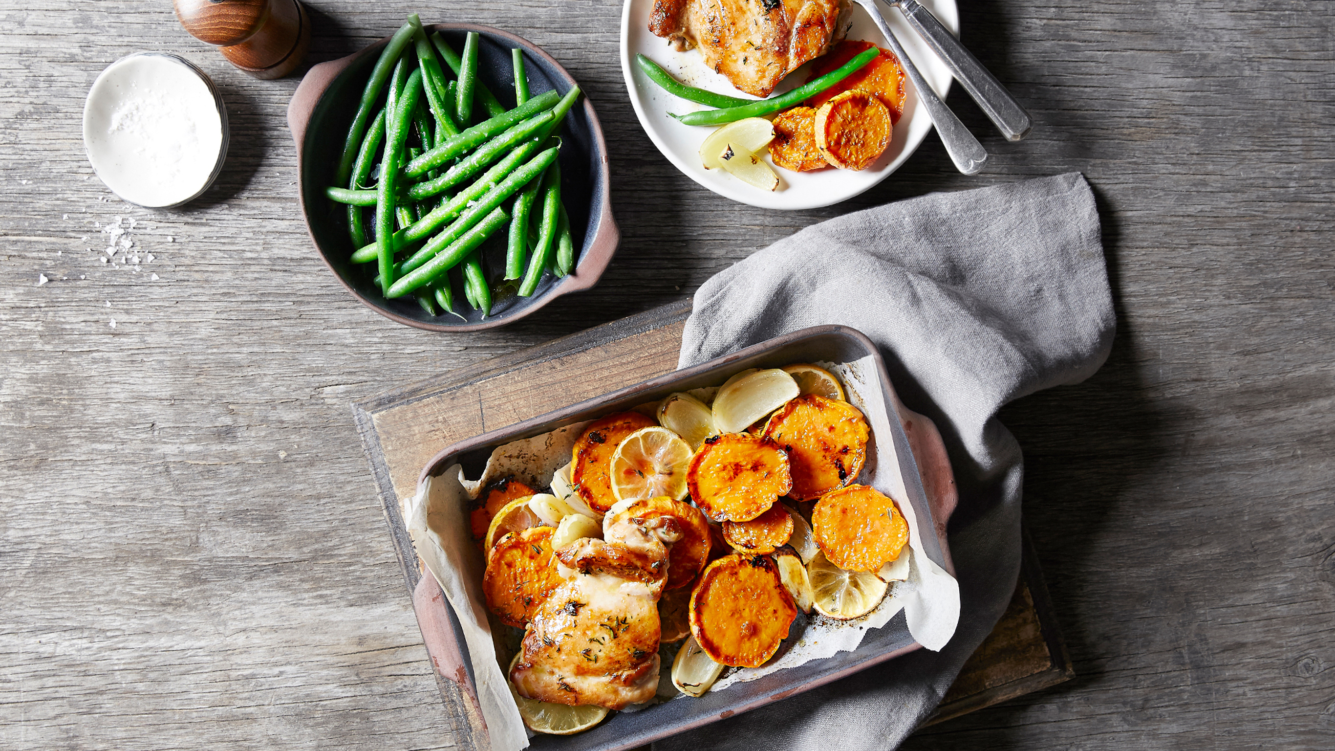 A tray of Centr's Lemon & Thyme Roast Chicken rests on a wooden board with a dish of green beans and a plate of chicken and beans on the side.