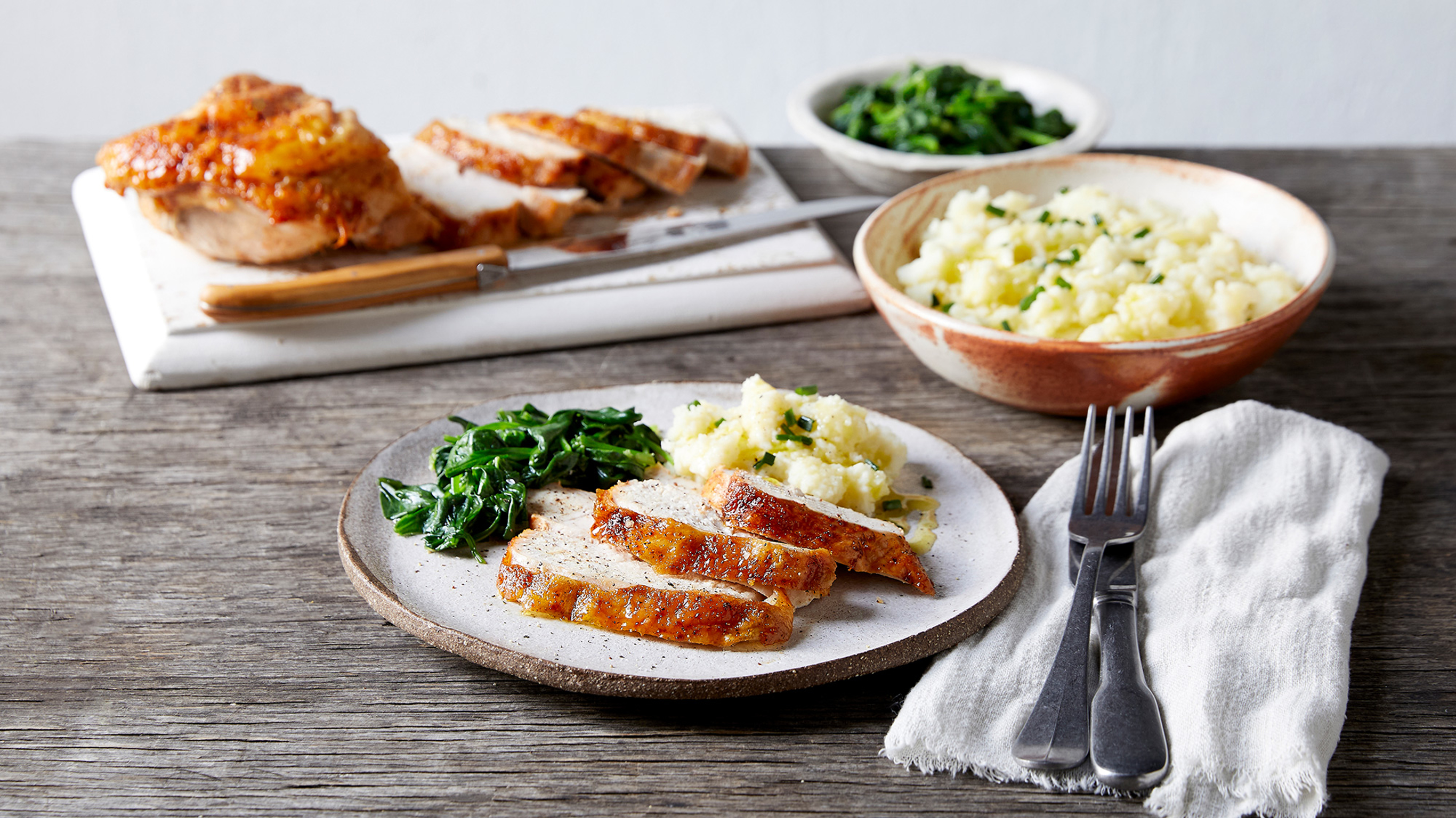 A plate of Centr's Roasted Turkey, Mashed Potatoes & Greens rests on rustic wooden table, knife and fork on a linen napkin, with extra potatoes and greens on the side.