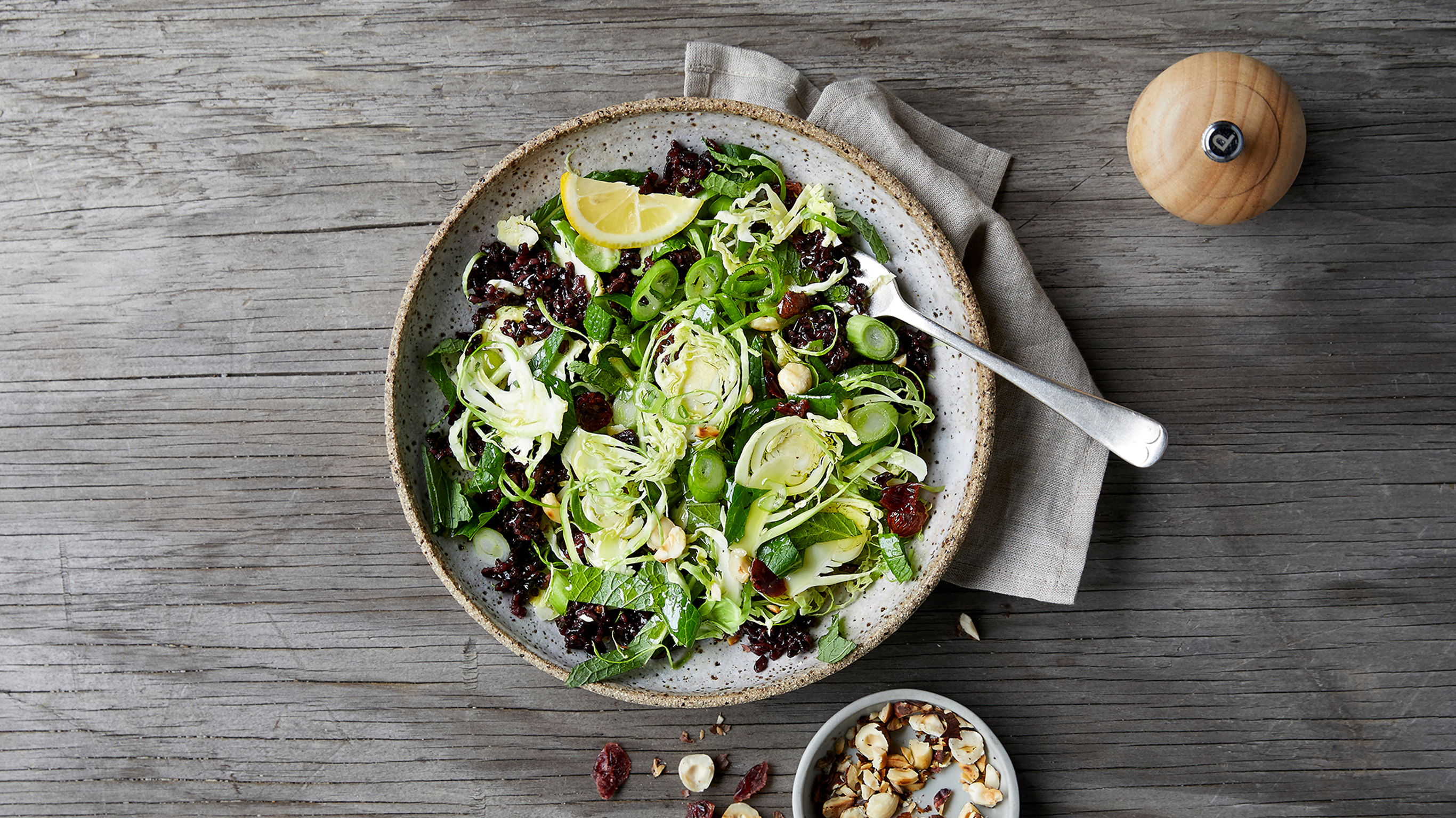 A plate of Centr's Shredded Brussel Sprouts Salad rests on a wooden table with a grey linen napkin tucked under the plate, and a pepper grind on the side.
