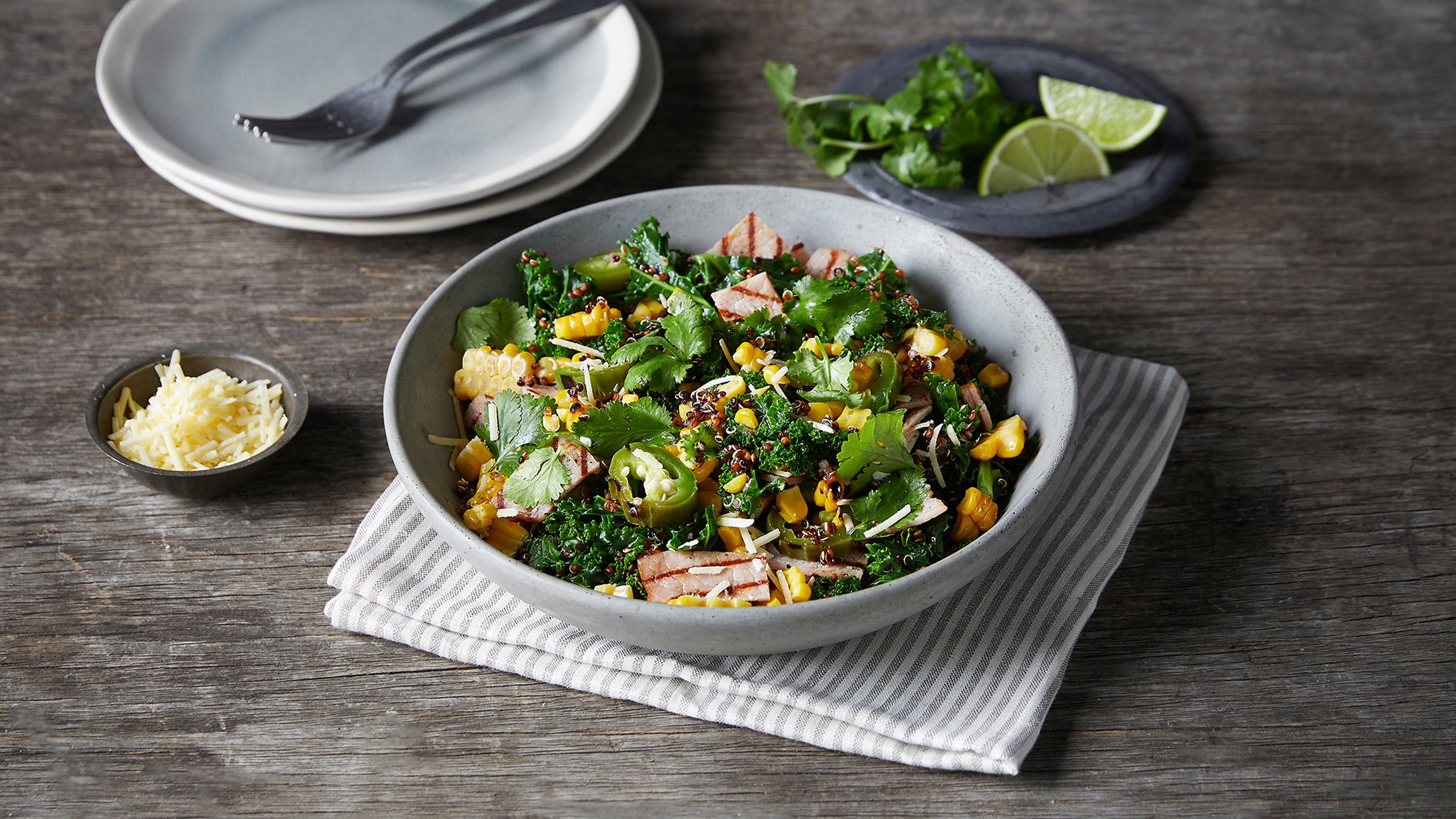 A plate of Charred Corn & Kale Salad with Bacon sits on a grey striped tea towel,  on a wooden table with small side dishes of grated parmesan, lime wedges and cilantro sprigs.