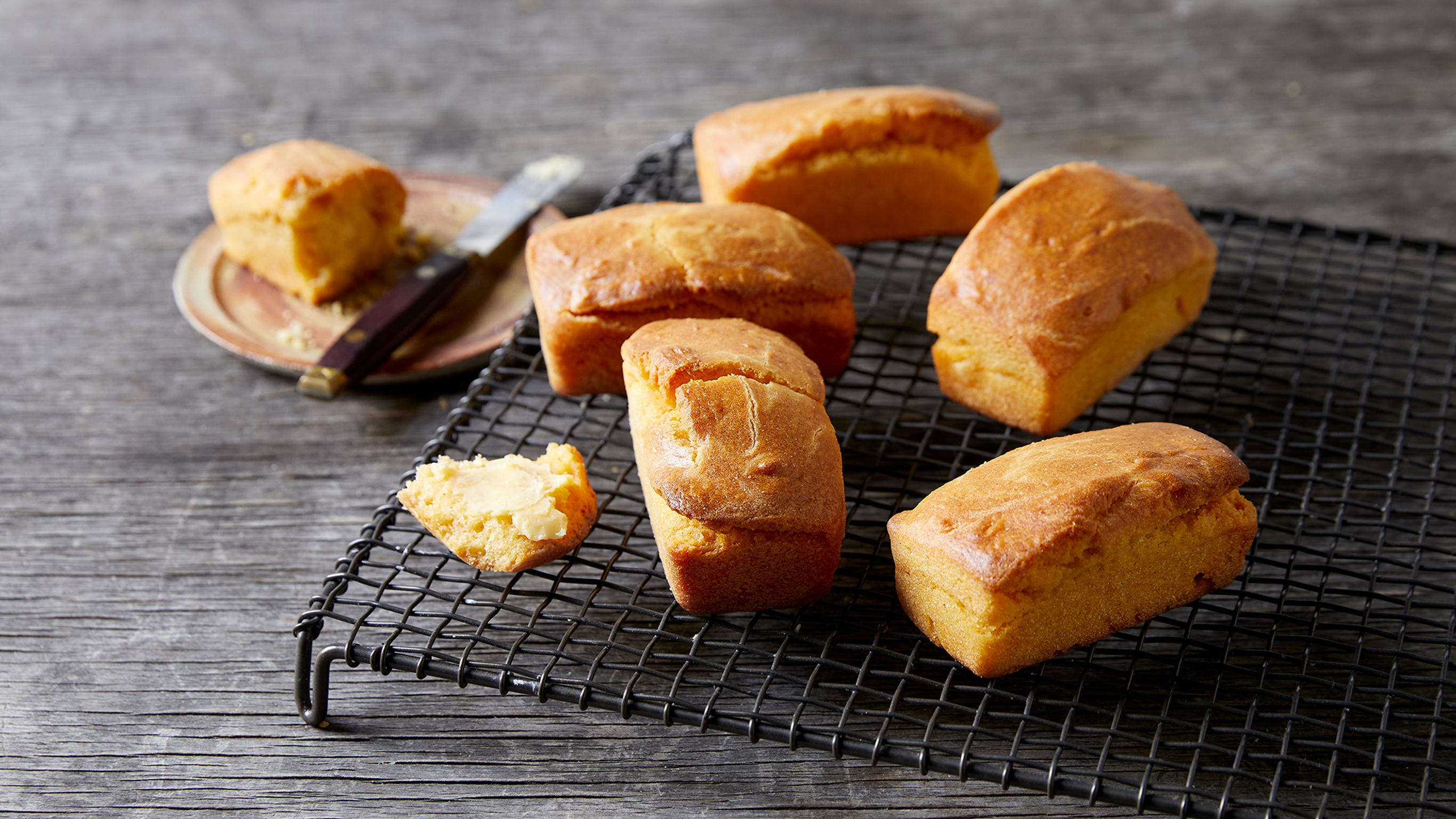 5 of Centr's mini corn muffins sit on a black cooling rack on a wooden table, with a cut muffin sitting on a plate to the side.