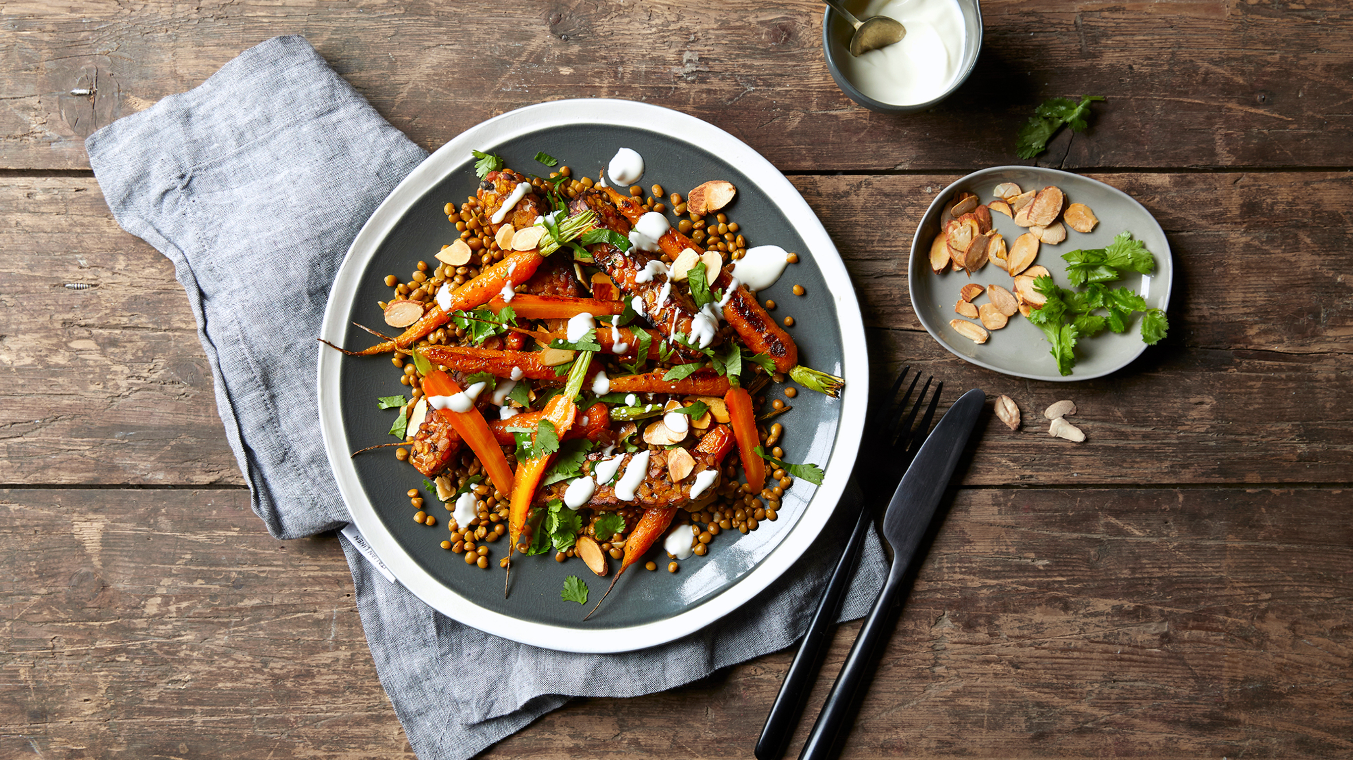 A plate of Centr's Roasted Carrot, Lentil & Almond Salad rests on a wooden table, with a grey linen napkin tucked underneath, silver knife and fork on the side.