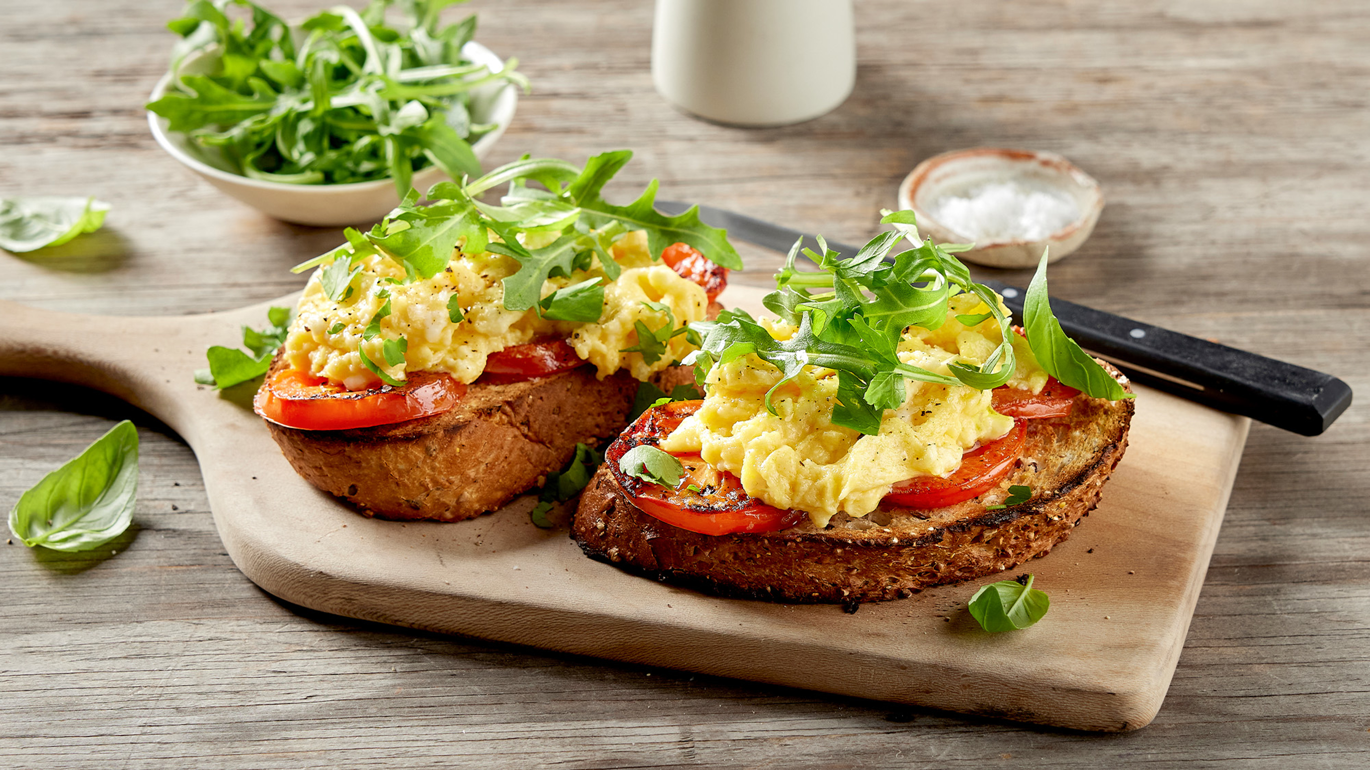Two slices of toast with Dan's Herby Egg Scramble Stack sit on a wooden serving board, with scattered basil leaves around and a small bowl of rocket leaves in the background.