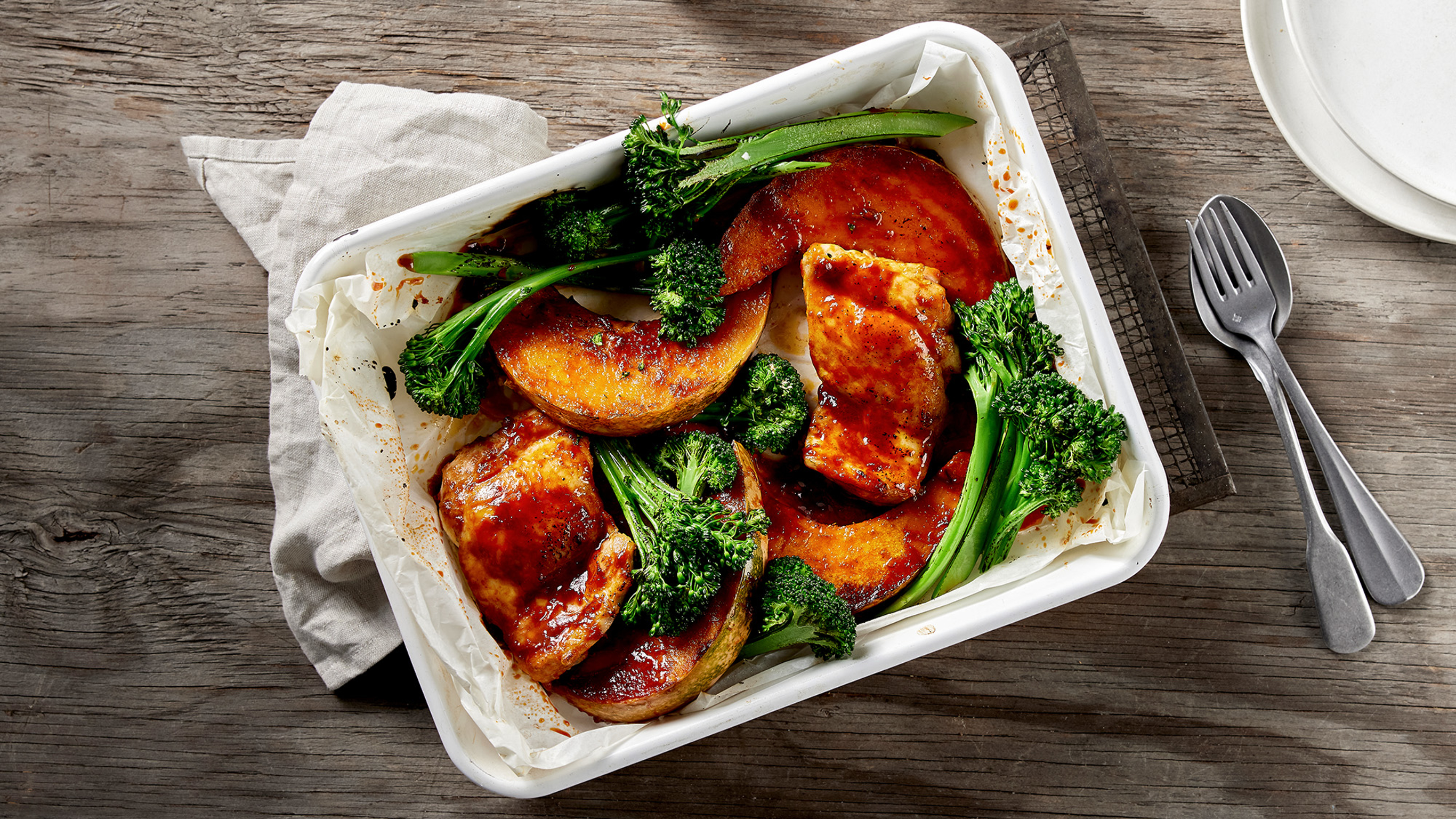 A tray of 6-ingredient Chilli Chicken & Squash Sheet-pan rests on a baking rack on a wooden table with a fork and spoon and plates on the side, ready for serving