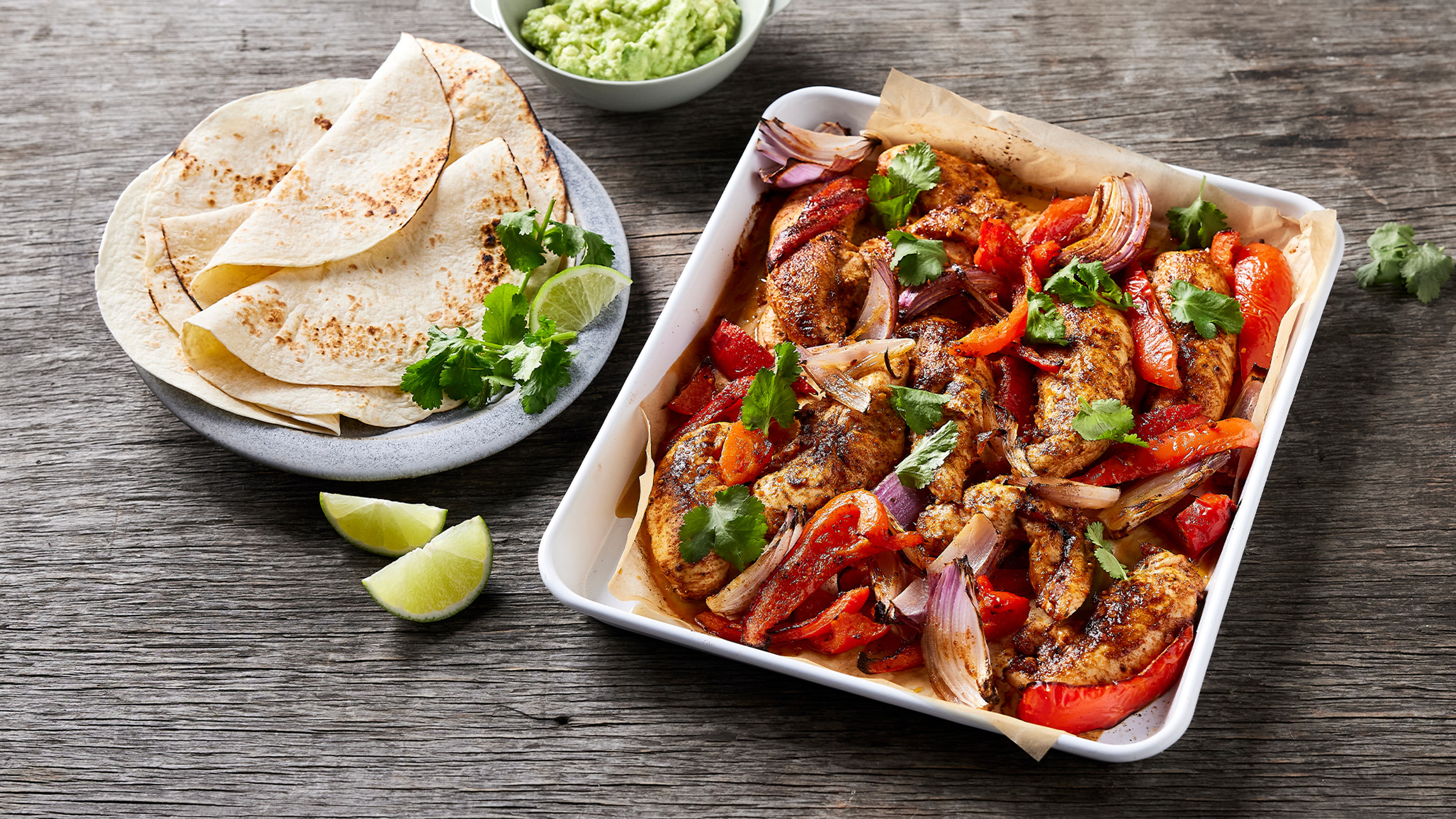 A tray of Centr's 6-ingredient Chicken Fajita Sheet-pan rests on a wooden table with a plate of tortillas and some lime quarters on the side.