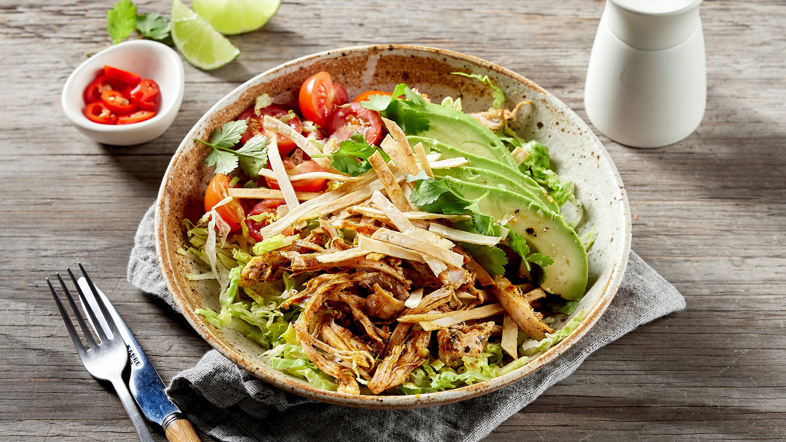 A bowl of Centr's 6-ingredient Crispy Chicken Taco Salad sits on a grey linen napkin on a wooden table, with lime quarters and slices of chilli in a bowl in the background.