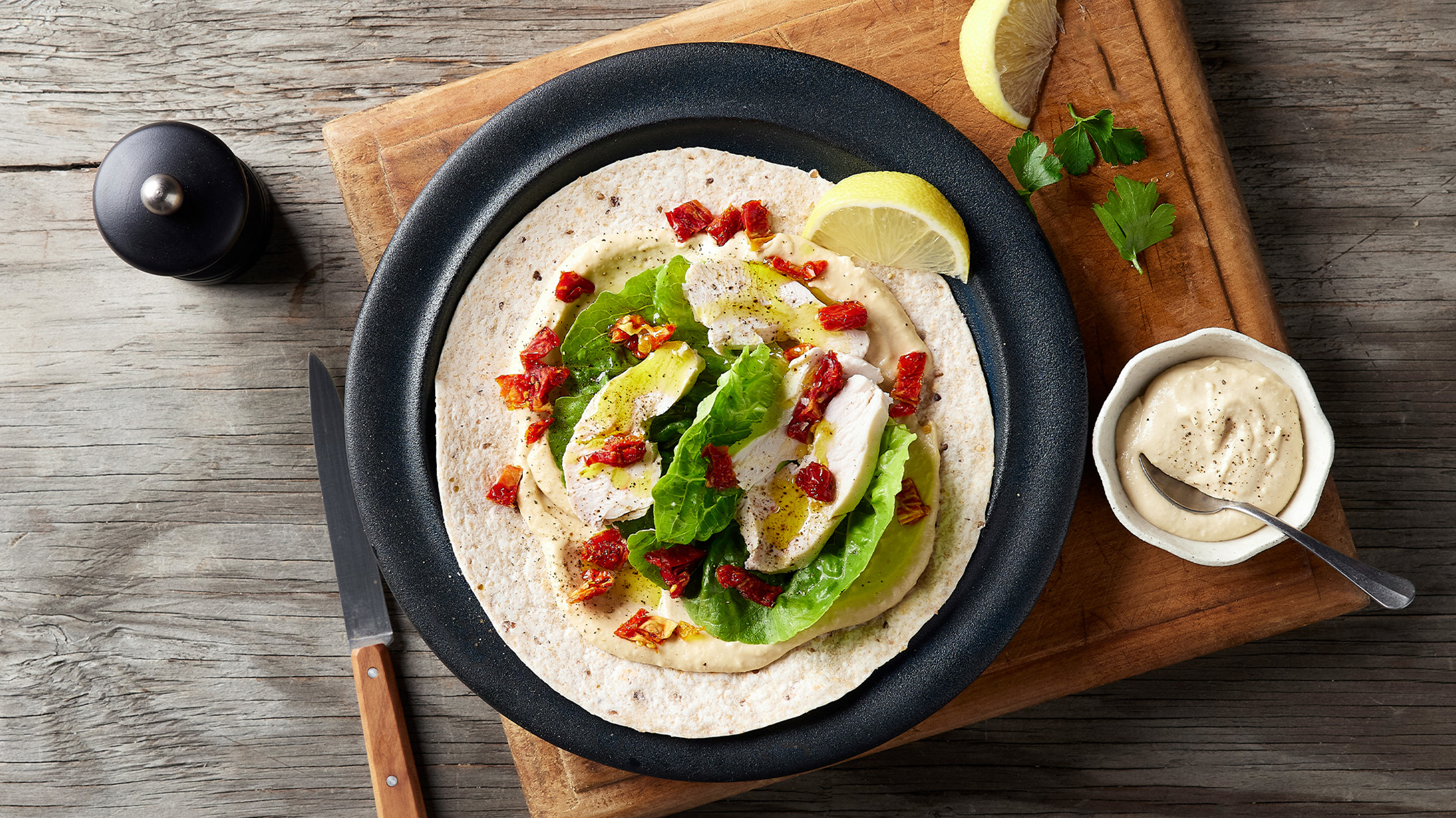 A tortilla piled with hummus lettuce, roast turkey, sundried tomatoes sits on a stone plate on a wooden board.