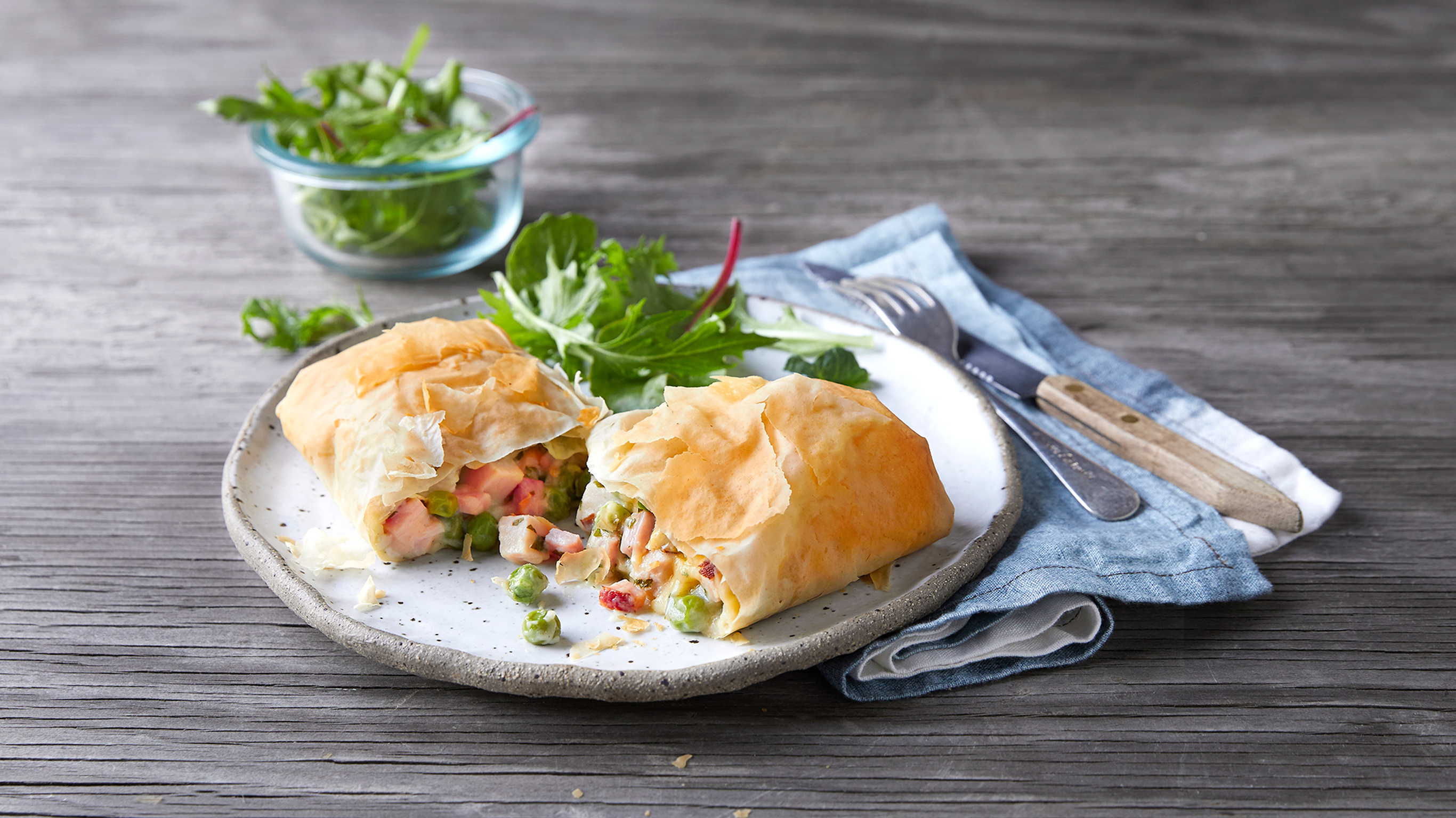 A  turkey & ham filo pie, cut in half, rests on a grey plate with a blue linen napkin, knife and fork on the side.
