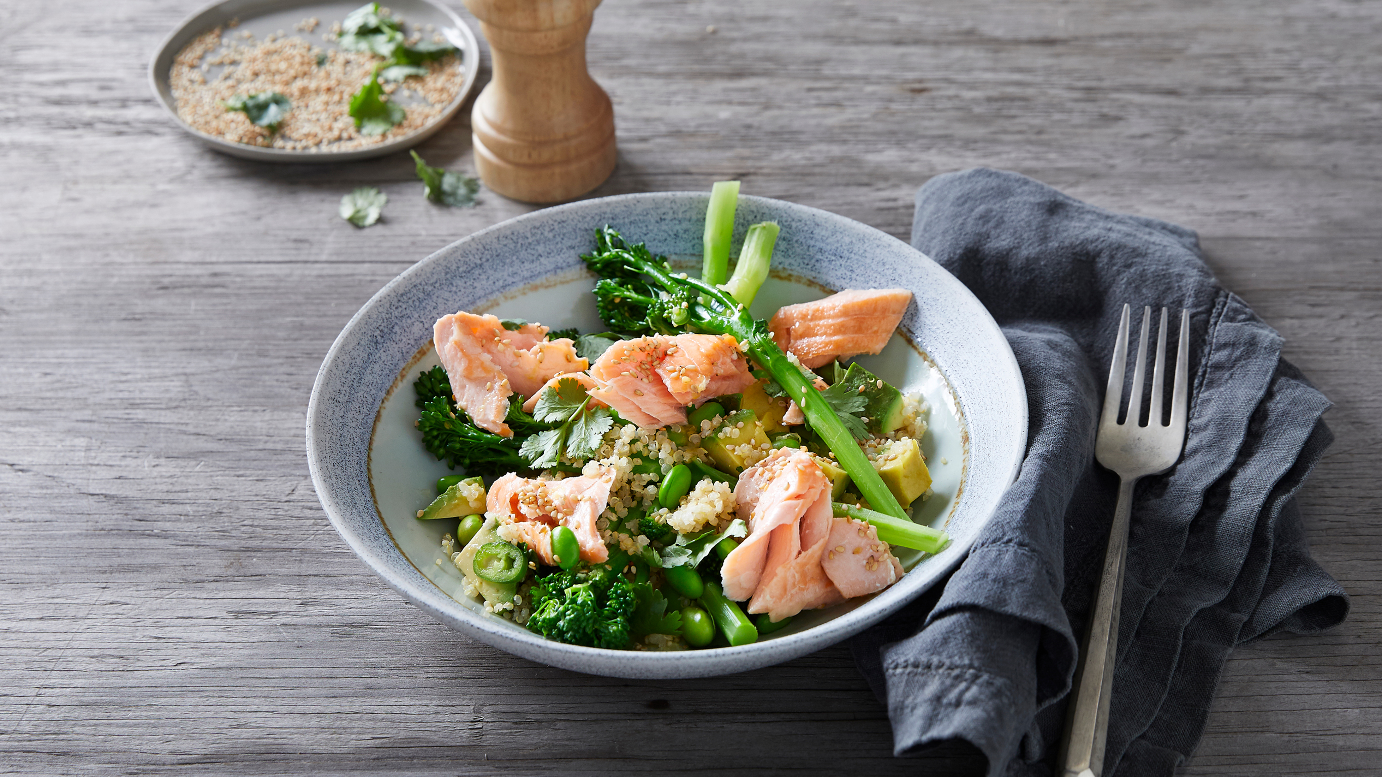 A bowl of Salmon, Edamame & Quinoa Salad rests on a wooden table, with a dark grey linen napkin and silver fork on the right hand side.