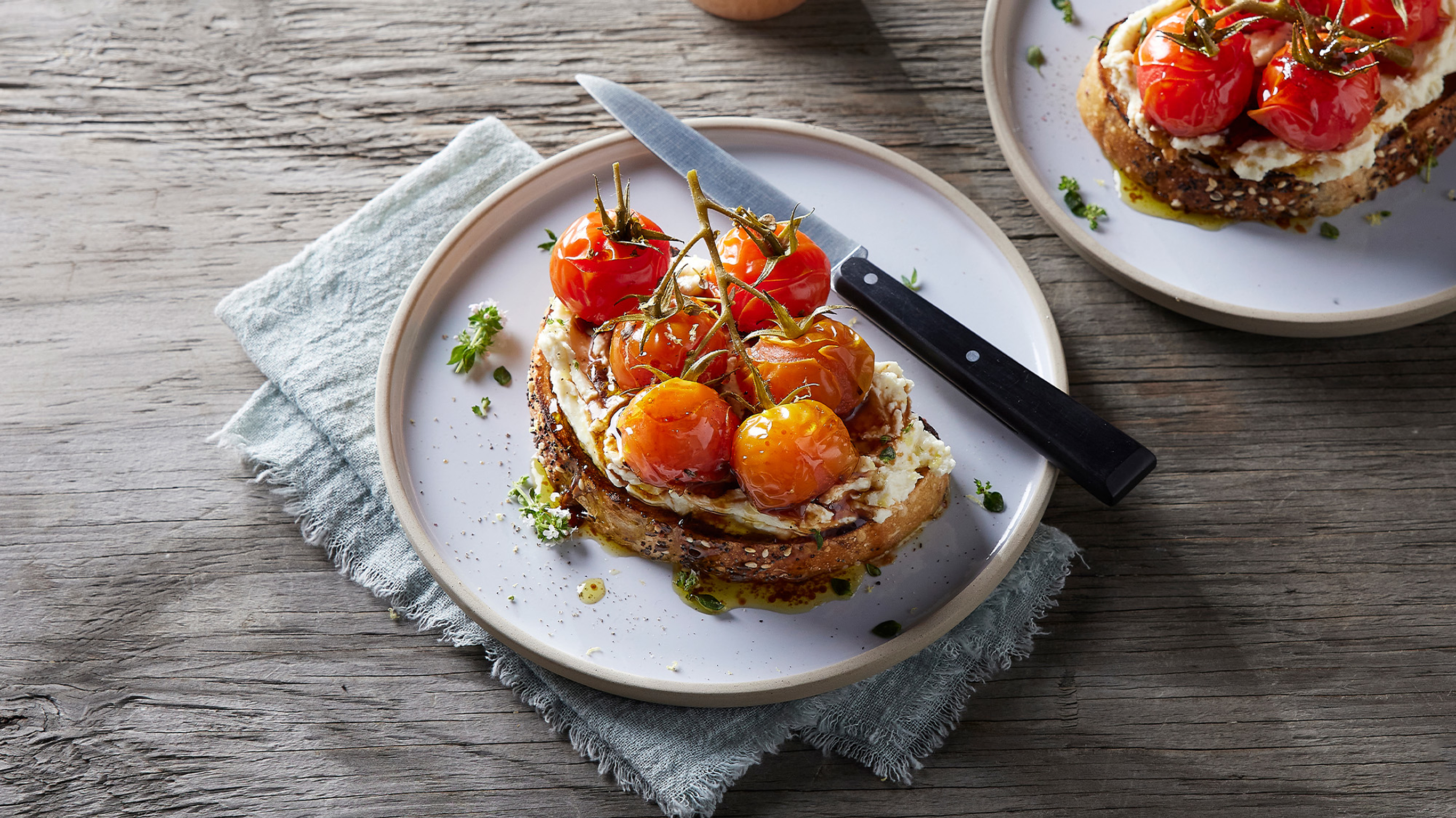 A slice of Centr's High protein cheese toast with roasted tomatoes sits on a round plate with a black-handled knife, and a second plate with toast to the side.