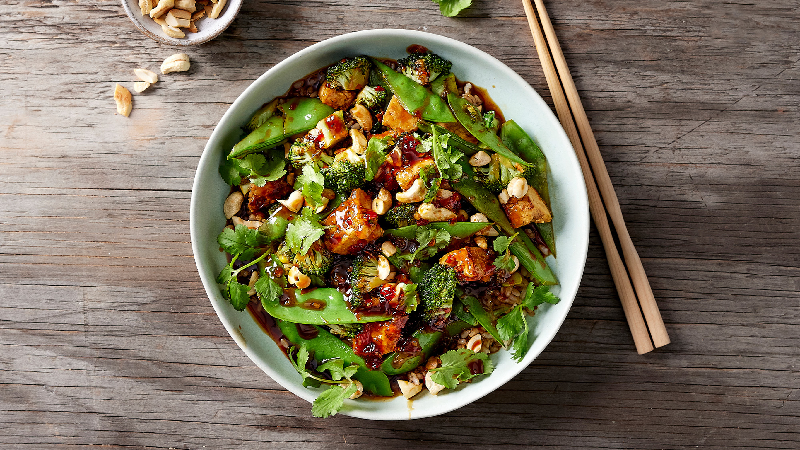 A bowl of Dan's Chilli Tofu & Cashew Stir Fry sits on a grey wooden table, with bamboo chopsticks and a small bowl of cashews on the side.
