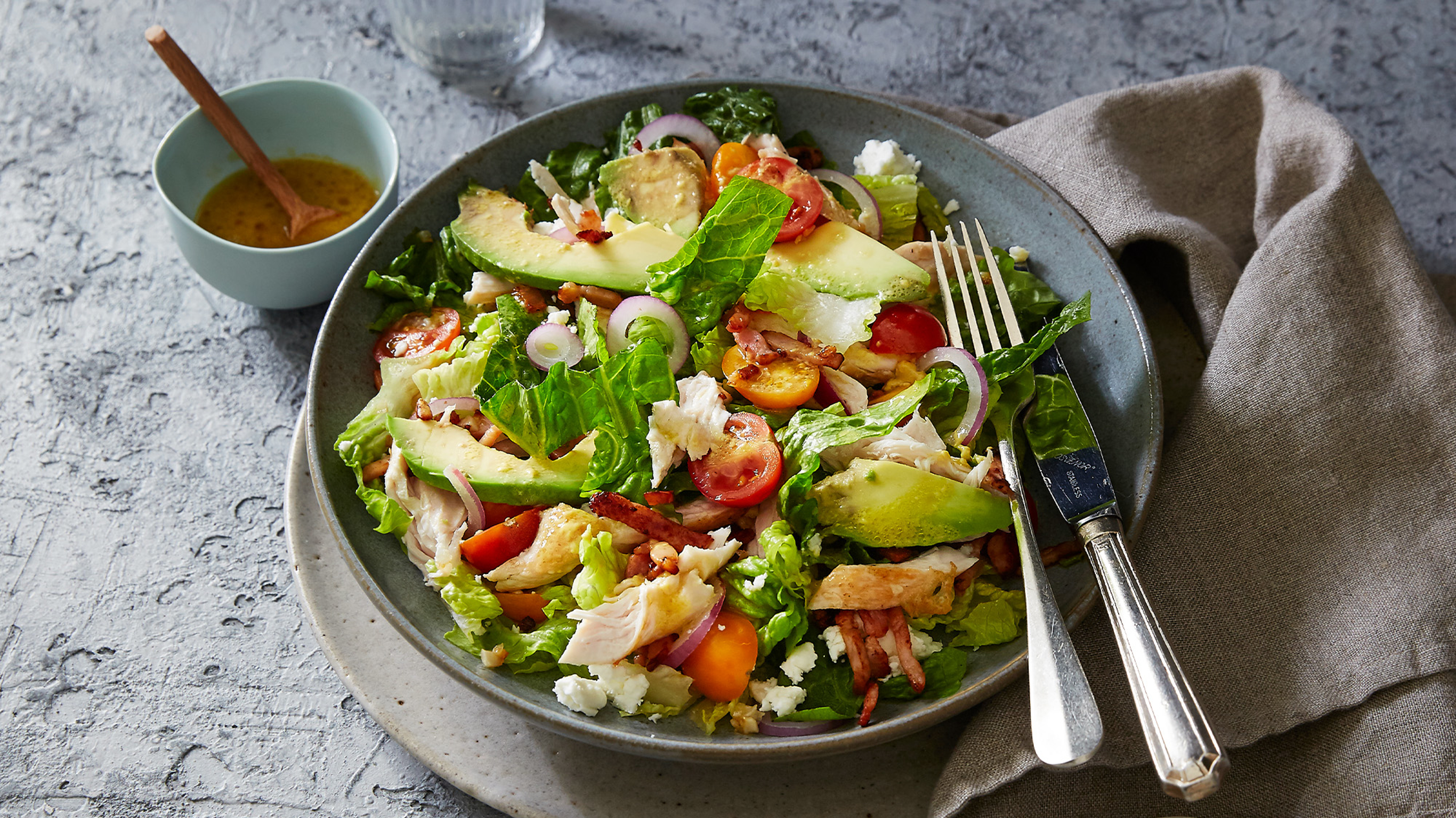 A serve of Chicken Cobb Salad with Honey Mustard Dressing in a grey bowl with silver knife and fork on the side rests on a table with a small bowl of honey mustard dressing to the side.