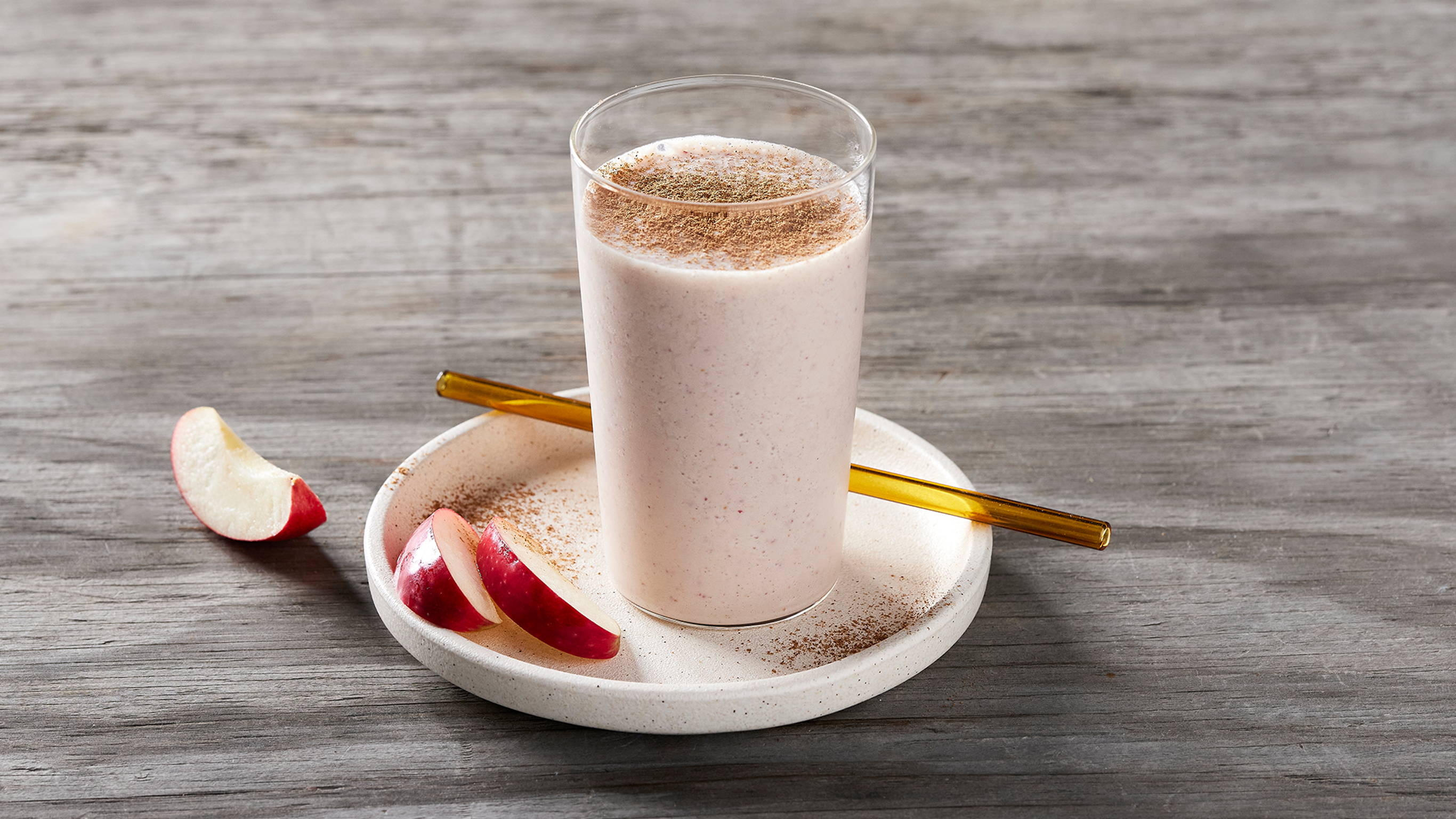 A tall glass of Centr's Caramel Apple Smoothie sits on a small round white stoneware plate, with slices of red-skinned apple on the side.
