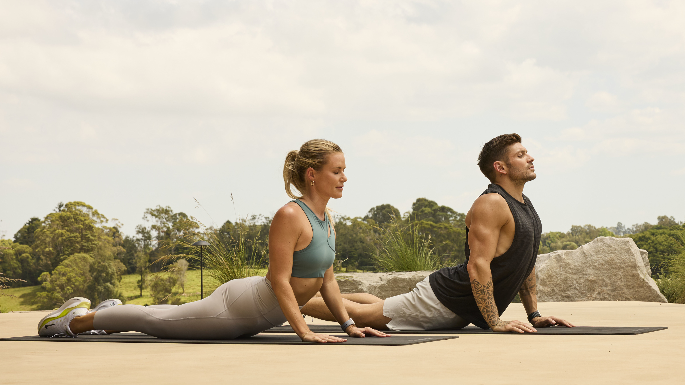 Alexz Parvi and Luke Zocchi perform cobra pose outdoors in a park setting.