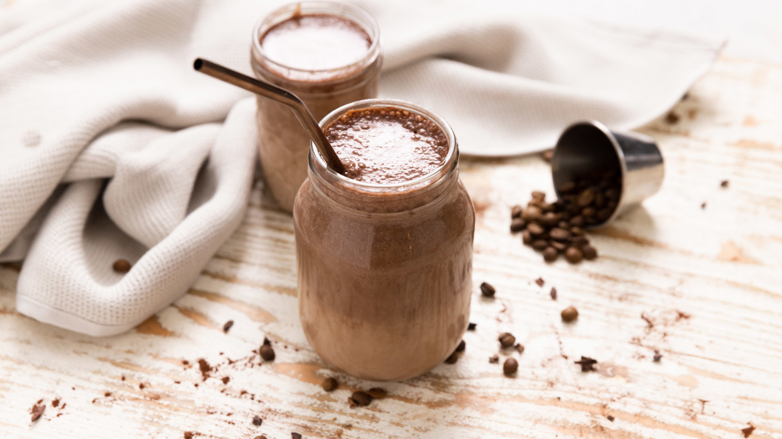 Two drinking jars of Centr's Coffee & Cacao Smoothie with bent, brown straws, rest on a wooden board, with coffee beans scattered around.