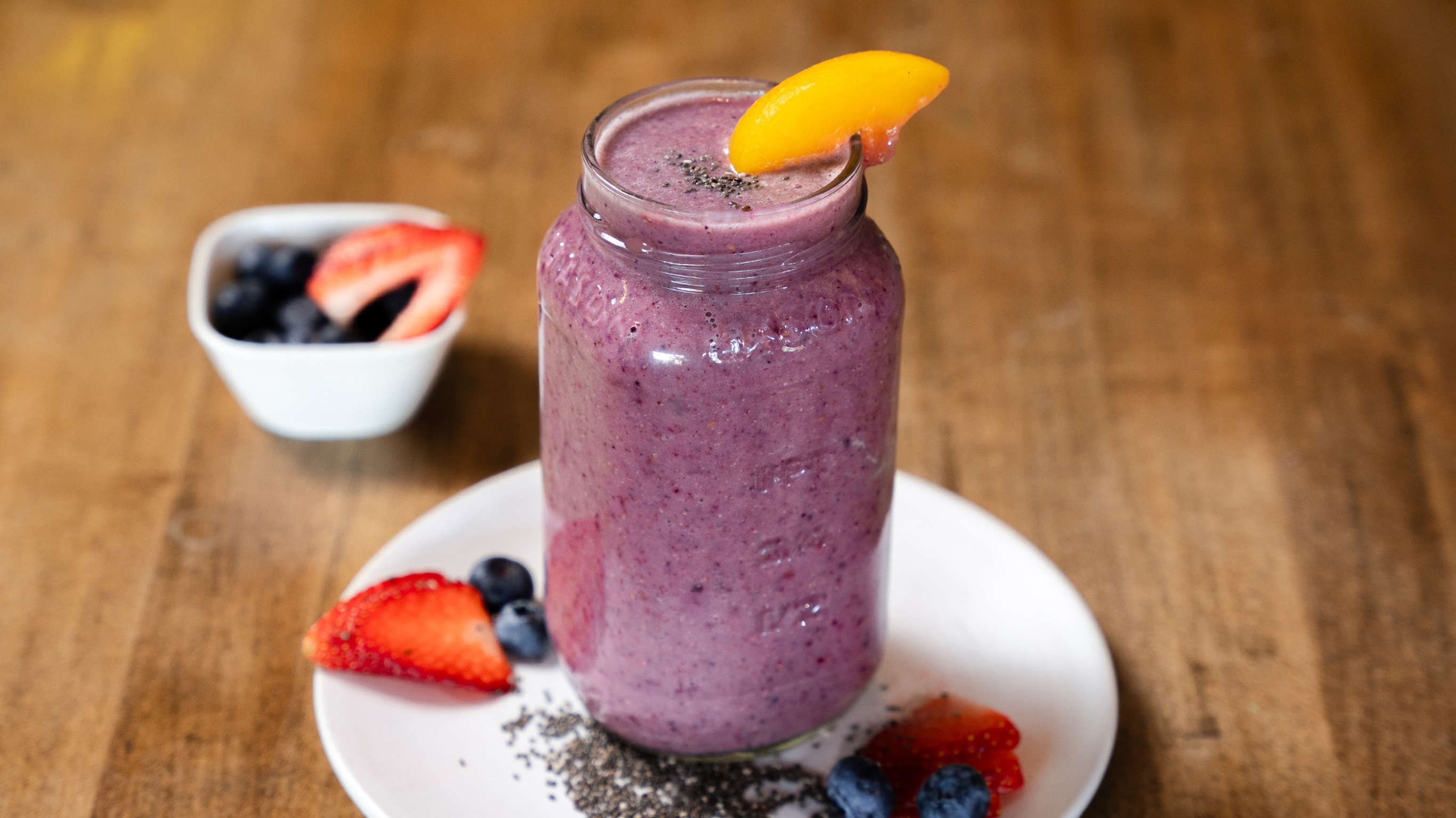 A drinking jar of Peach Almond Berry Reset Smoothie, decorated with a peach slice, sits on a white plate with slices of strawberrry, and blueberries scattered around.