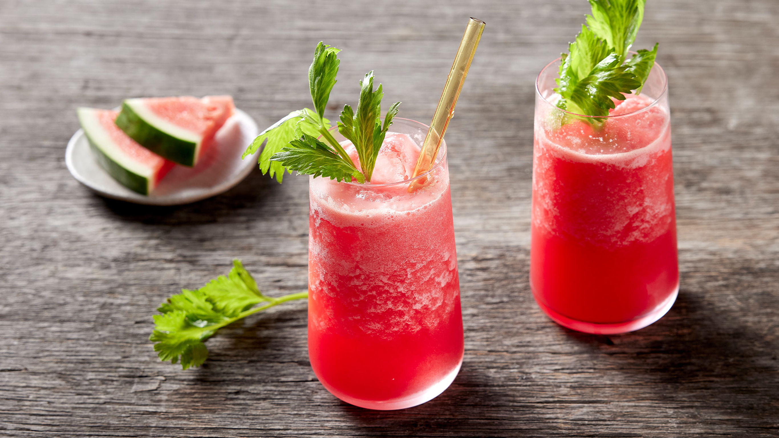 Two tall glasses of pink coloured Cleansing Celery & Watermelon Juice sit on a wooden table with a stick of celery and slices of watermelon behind.