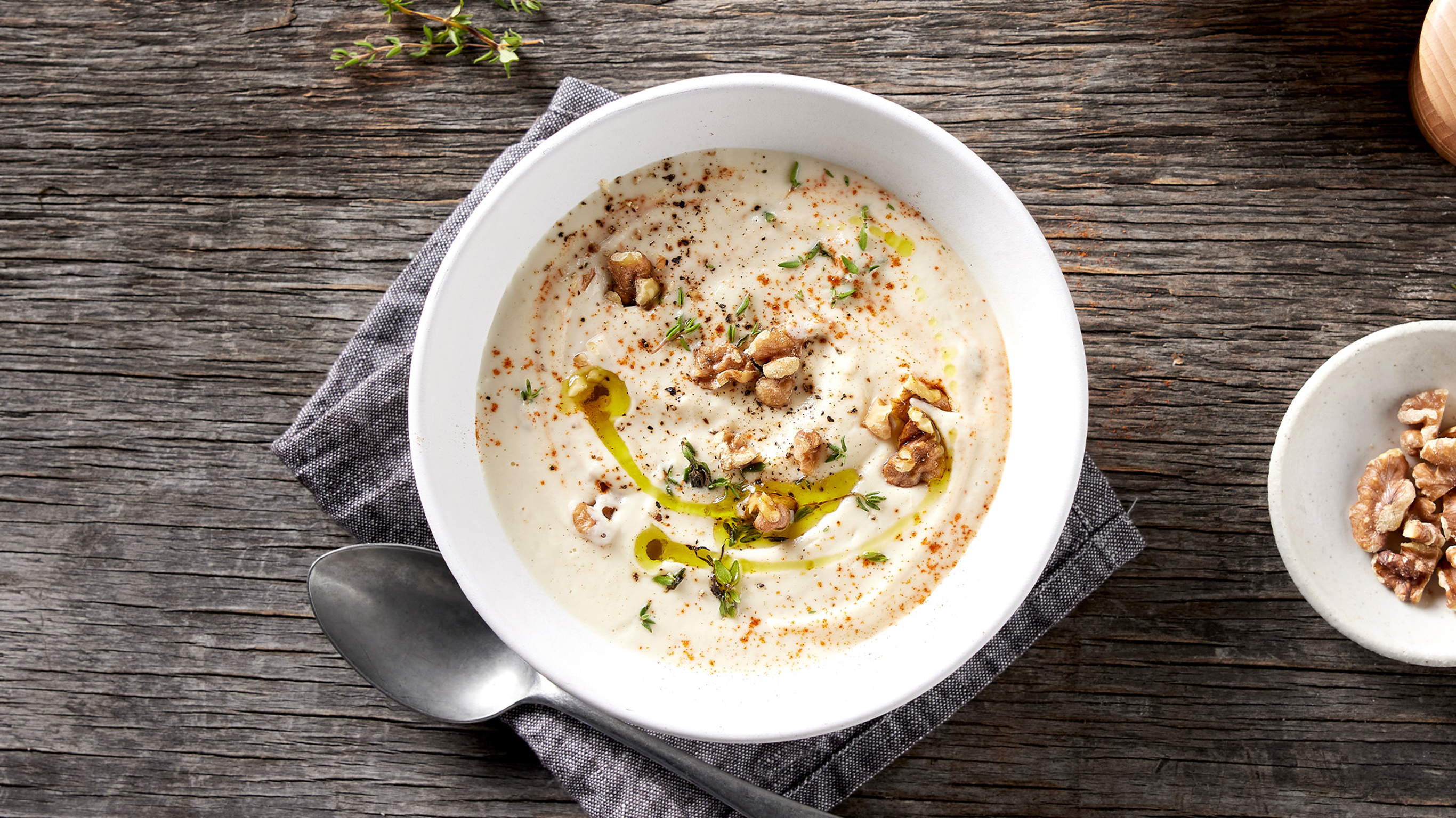 A white bowl of Creamy Cauliflower & Thyme Soup rests on a grey cotton napkin, with a spoon on the side ready for eating.