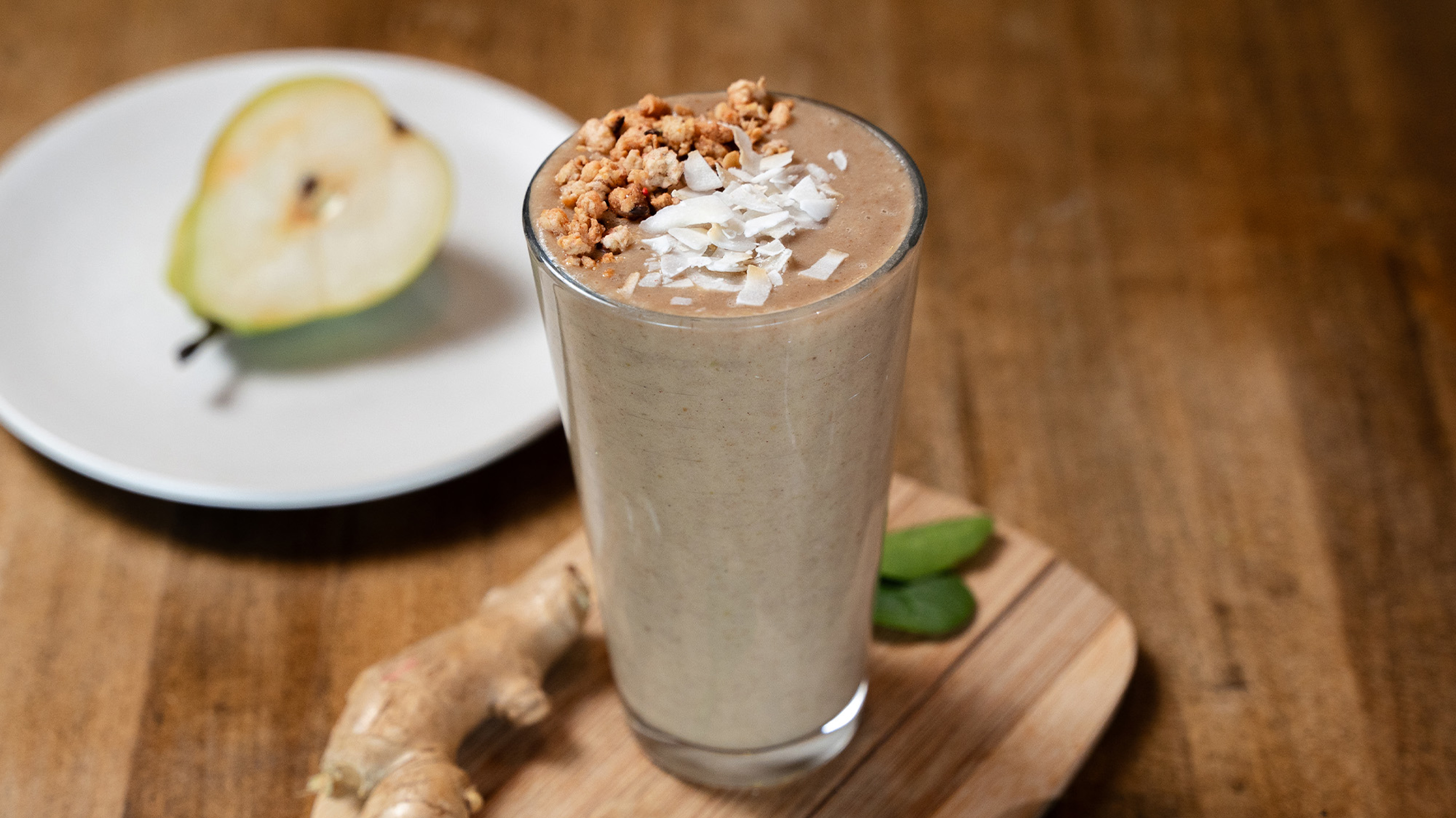 A tall glass of Centr's Ginger Honey Pear Reset Smoothie sits on a wooden board, with ginger on the side and half a pear on a plate behind.