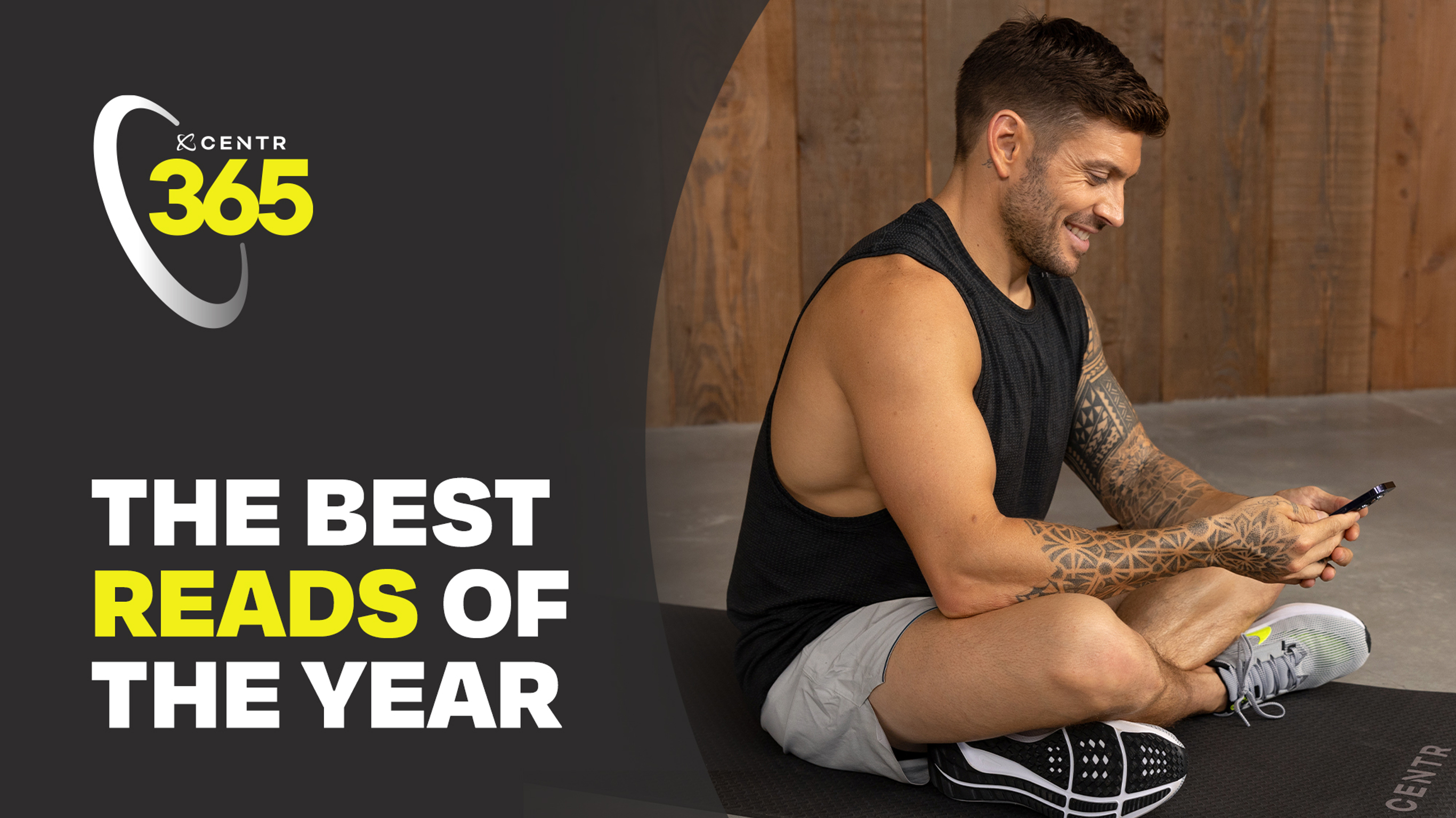 Centr trainer Luke Zocchi sits crossed legged on a yoga mat, smiling while reading an article on his phone.