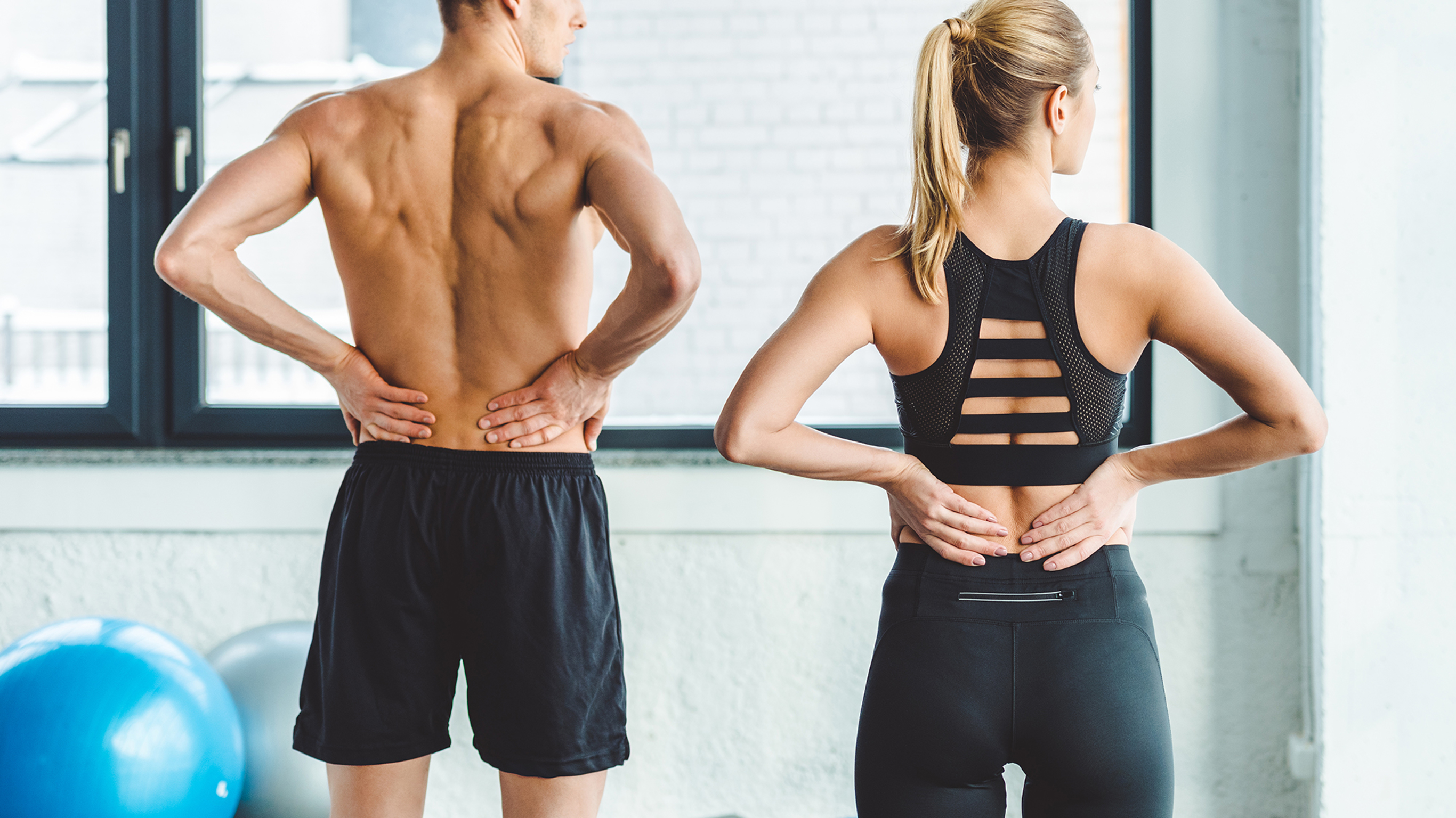 A man and a woman, both in gym wear with their backs to the camera, and have both hands placed on their lumbar back.