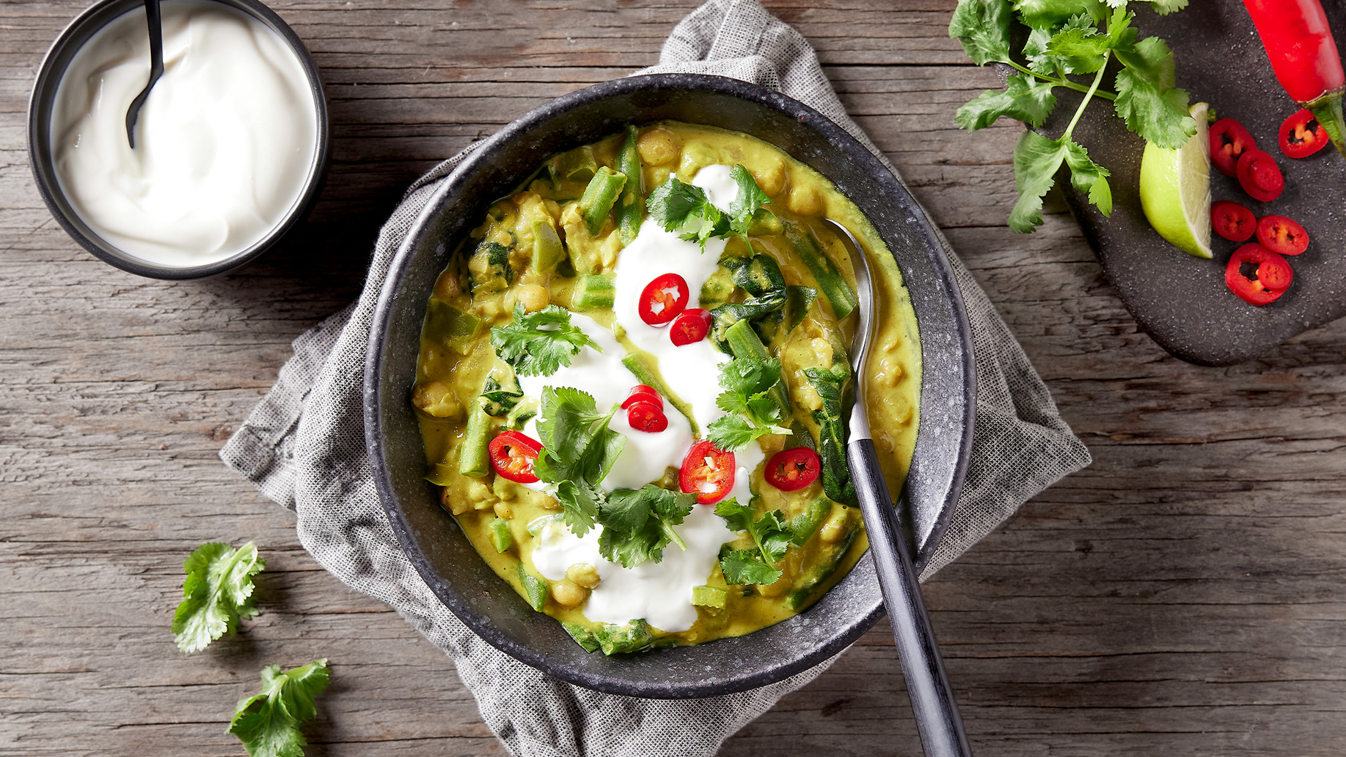 A bowl of Centr's Chickpea Coconut Dahl rest on a grey linen napkin on a wooden table, with black-handled spoon, ready for eating.