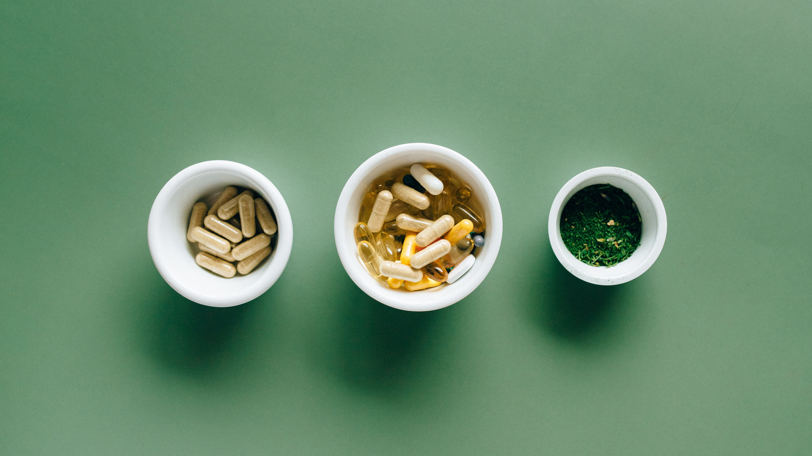 Three different sized white bowls holding supplements rest in a mid-green background.