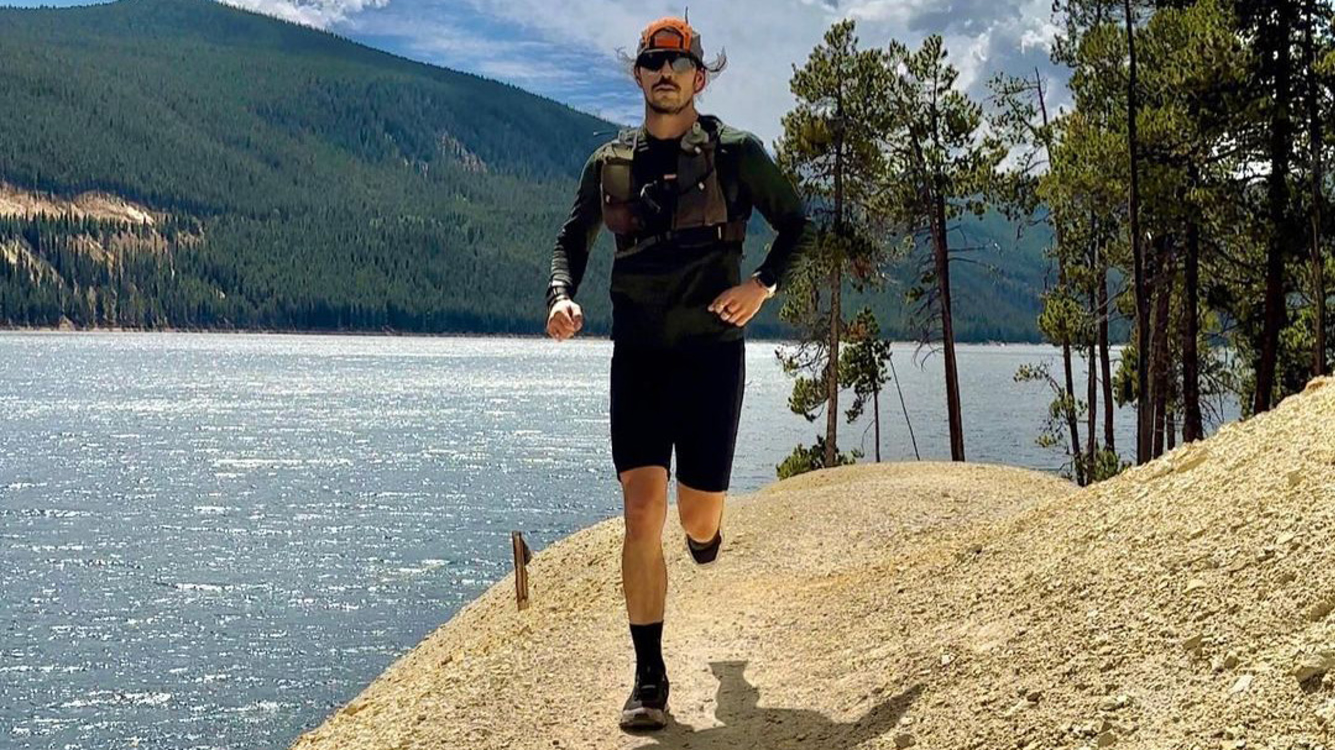 Dan Churchil, runs along a path on a gravel hill,  beside a mountain lake,  under a  blue, cloud-filled sky.