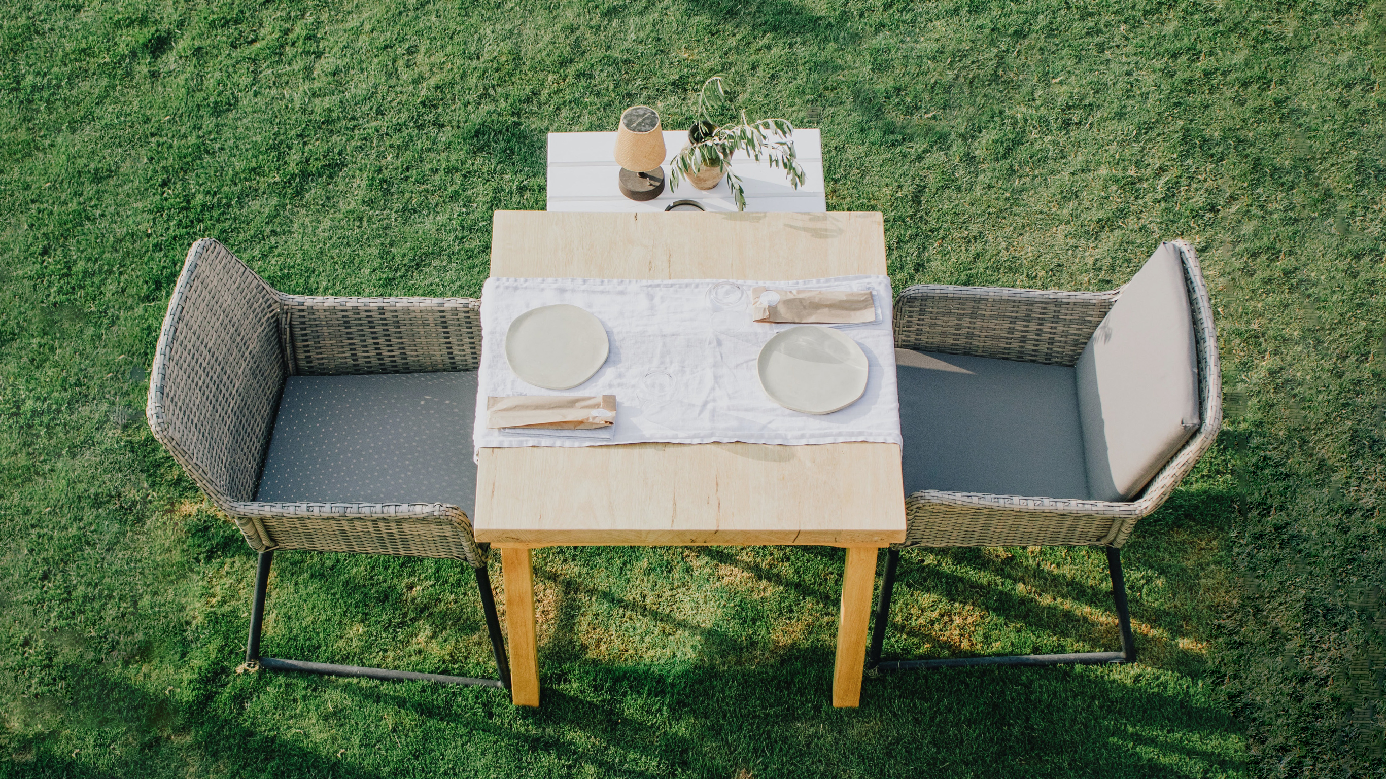 An overhead photo of a small square table, set with plates and cutlery in brown paper bags, for two, with two wicker chairs, outside on green lawn.