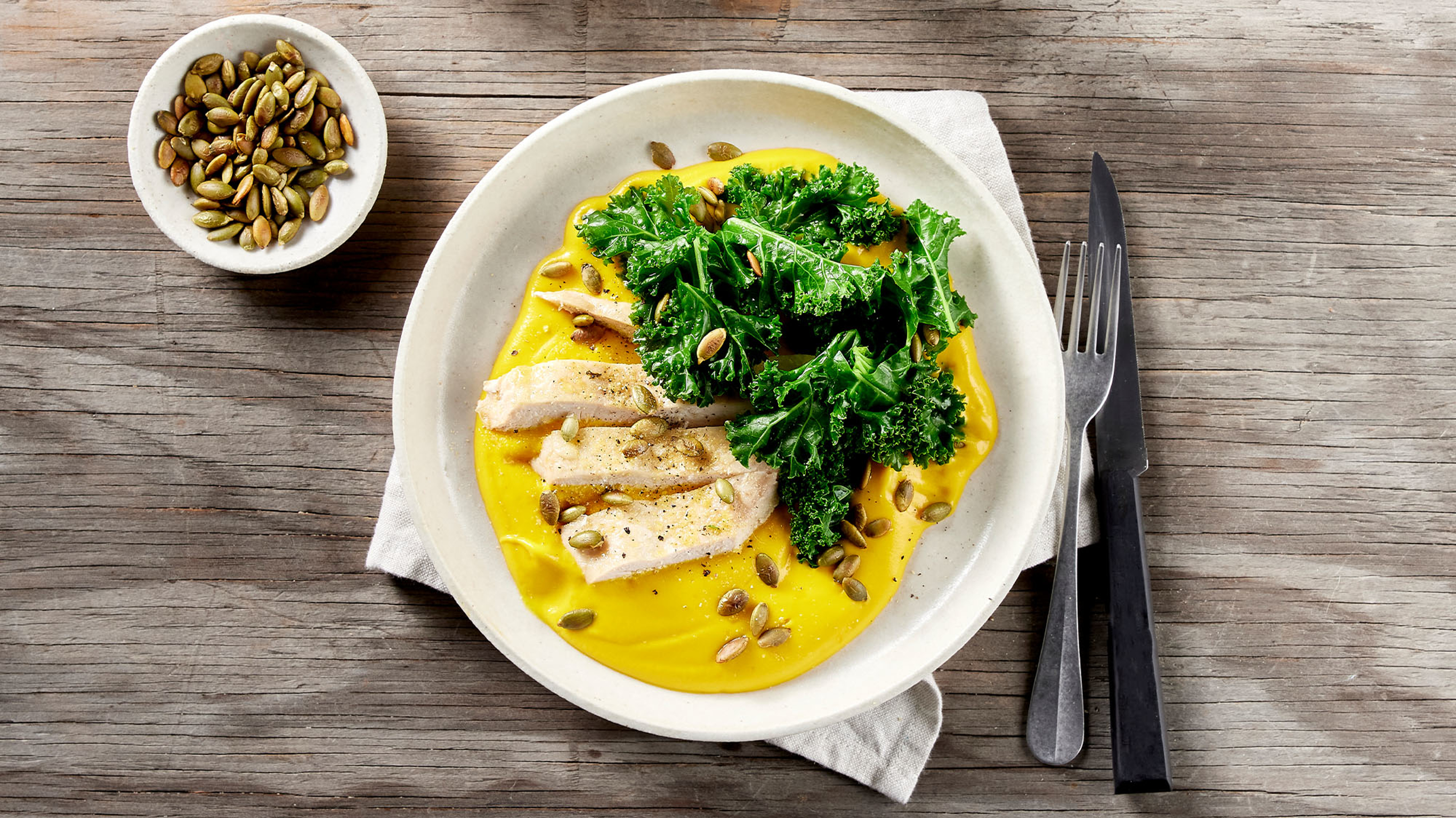 A bowl of Centr's 6-ingredient garlic chicken with sweet potato and kale sits on a white linen napkin, knife and fork to the side, ready for eating.