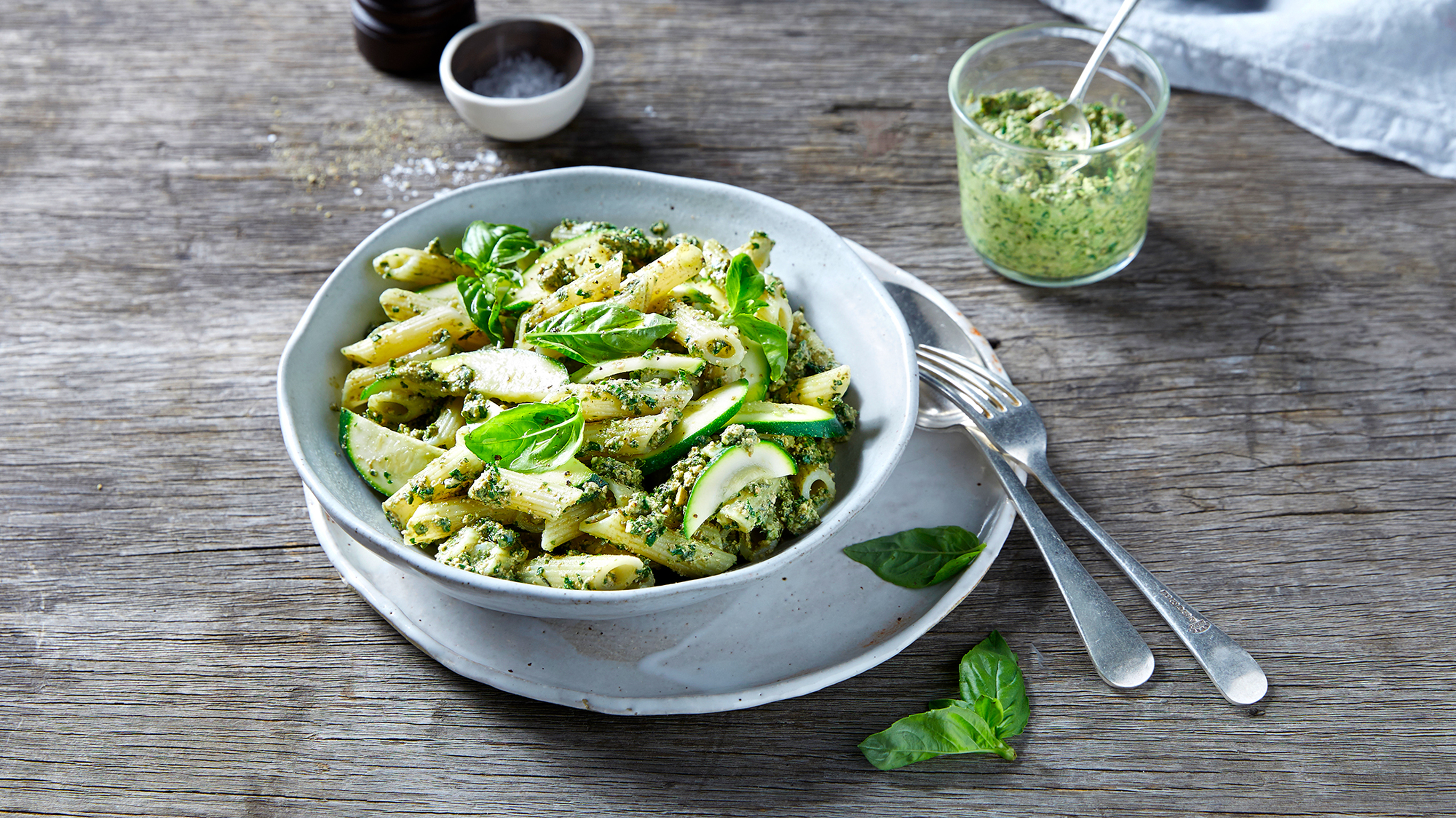 A bowl of pasta with kale & cashew pesto pasta sits on a plate, with a jar of extra kale & cashew pesto on the side.