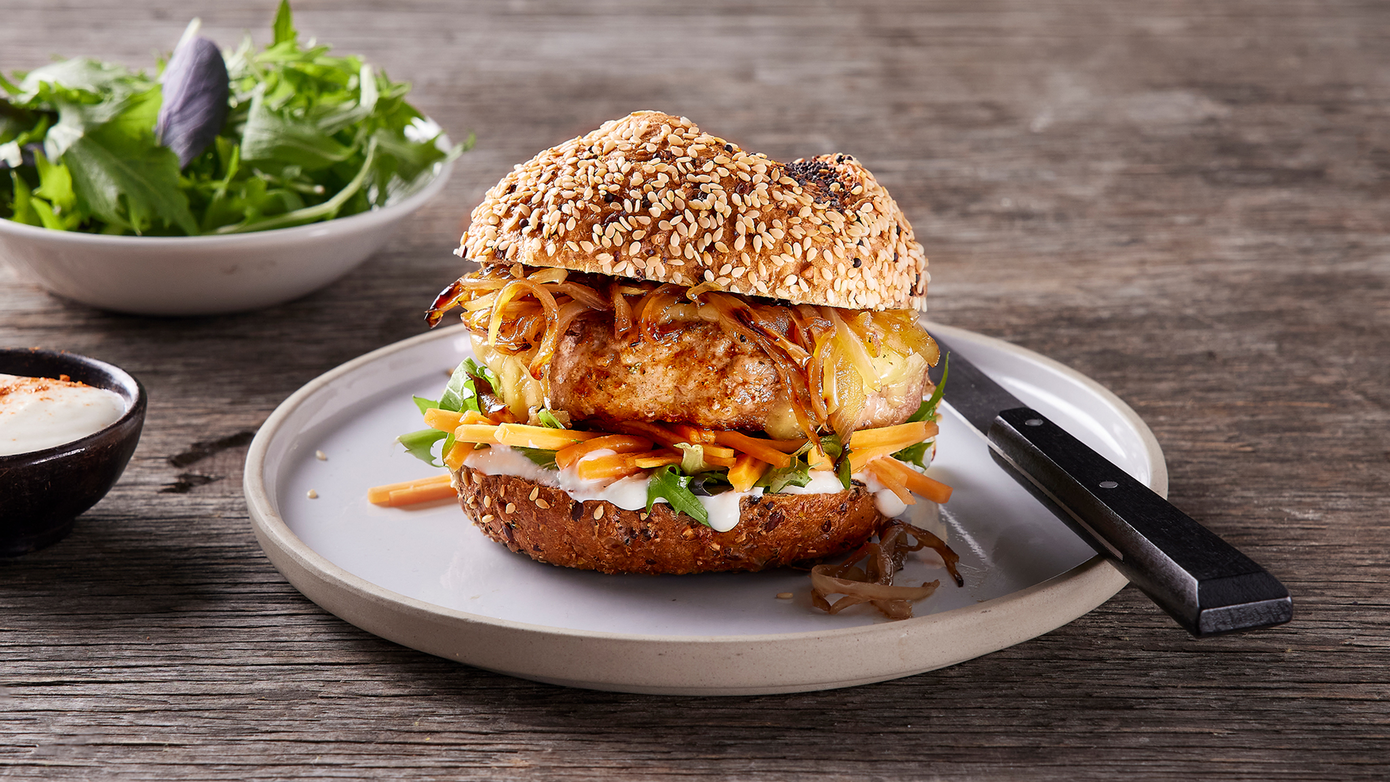 A toasted sesame seed bun with a Turkey burger patty, topped with caramelised onions sits on a white plate, ready for eating.