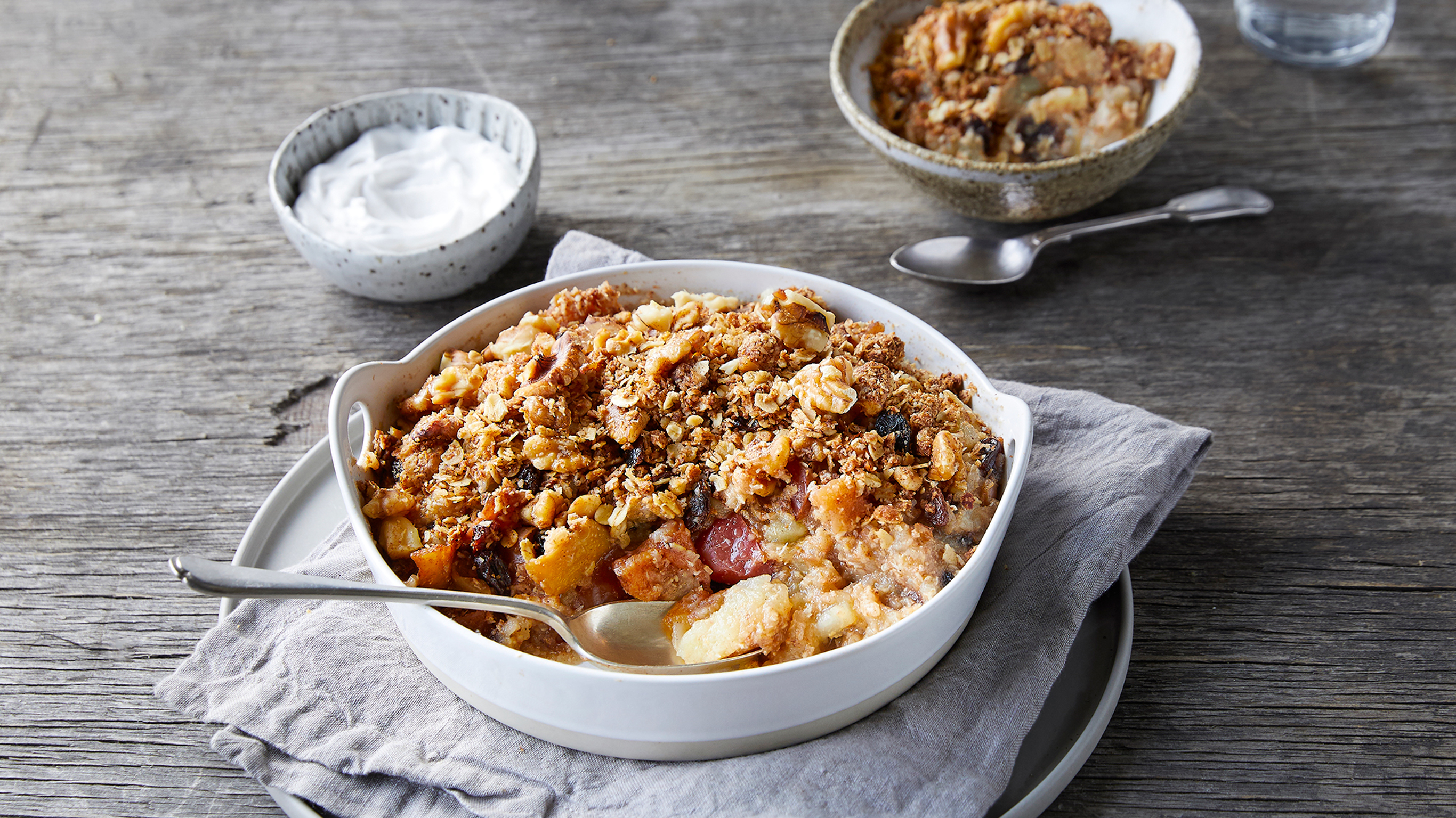 A baked apple breakfast crumble sitting on a table with yoghurt ready to eat.