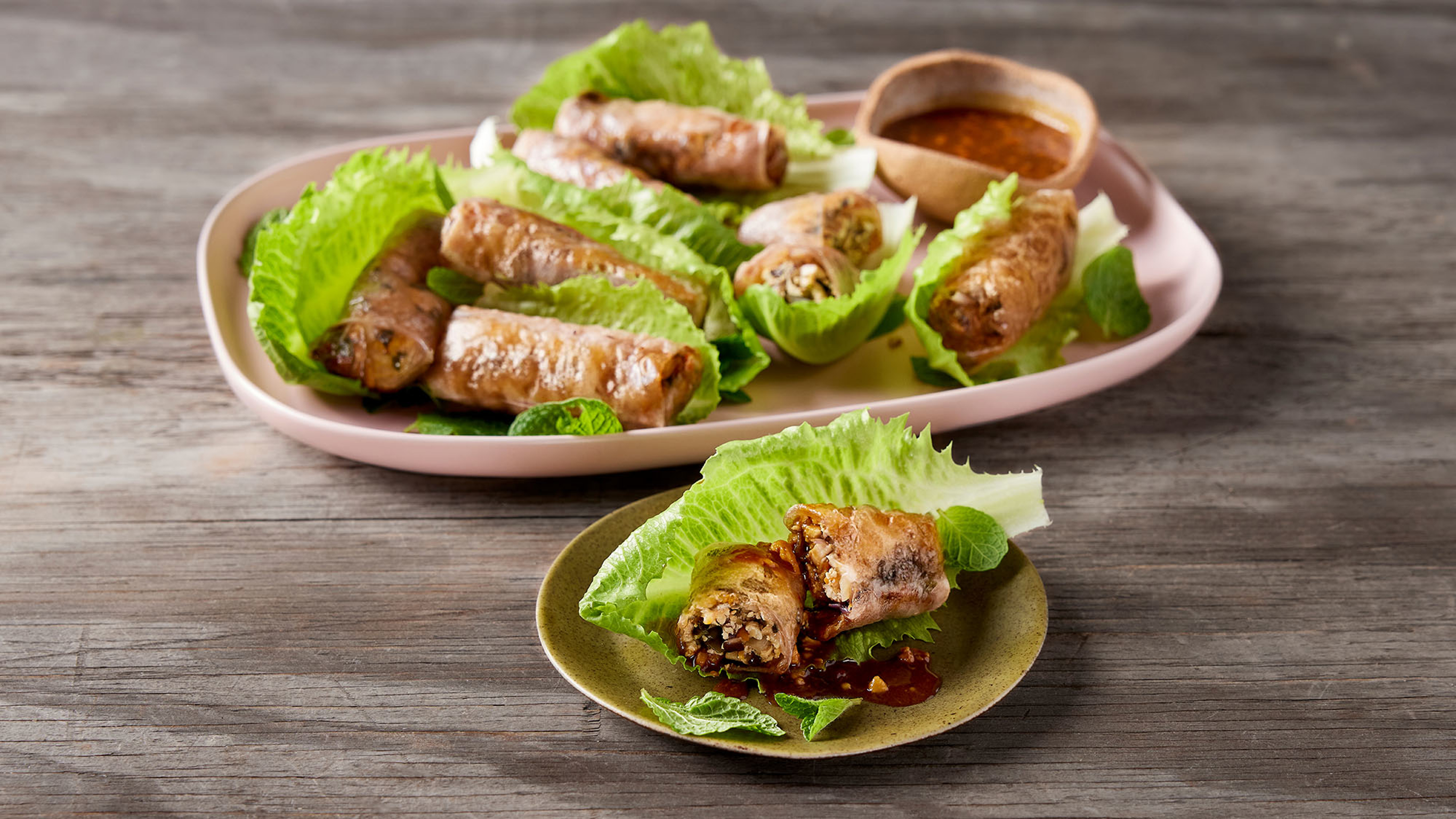 A serving platter of tofu & mushroom crispy rice paper rolls with lettuce leaves and dipping sauce, rests on a wooden table, with a serve on a small plate, ready for eating.