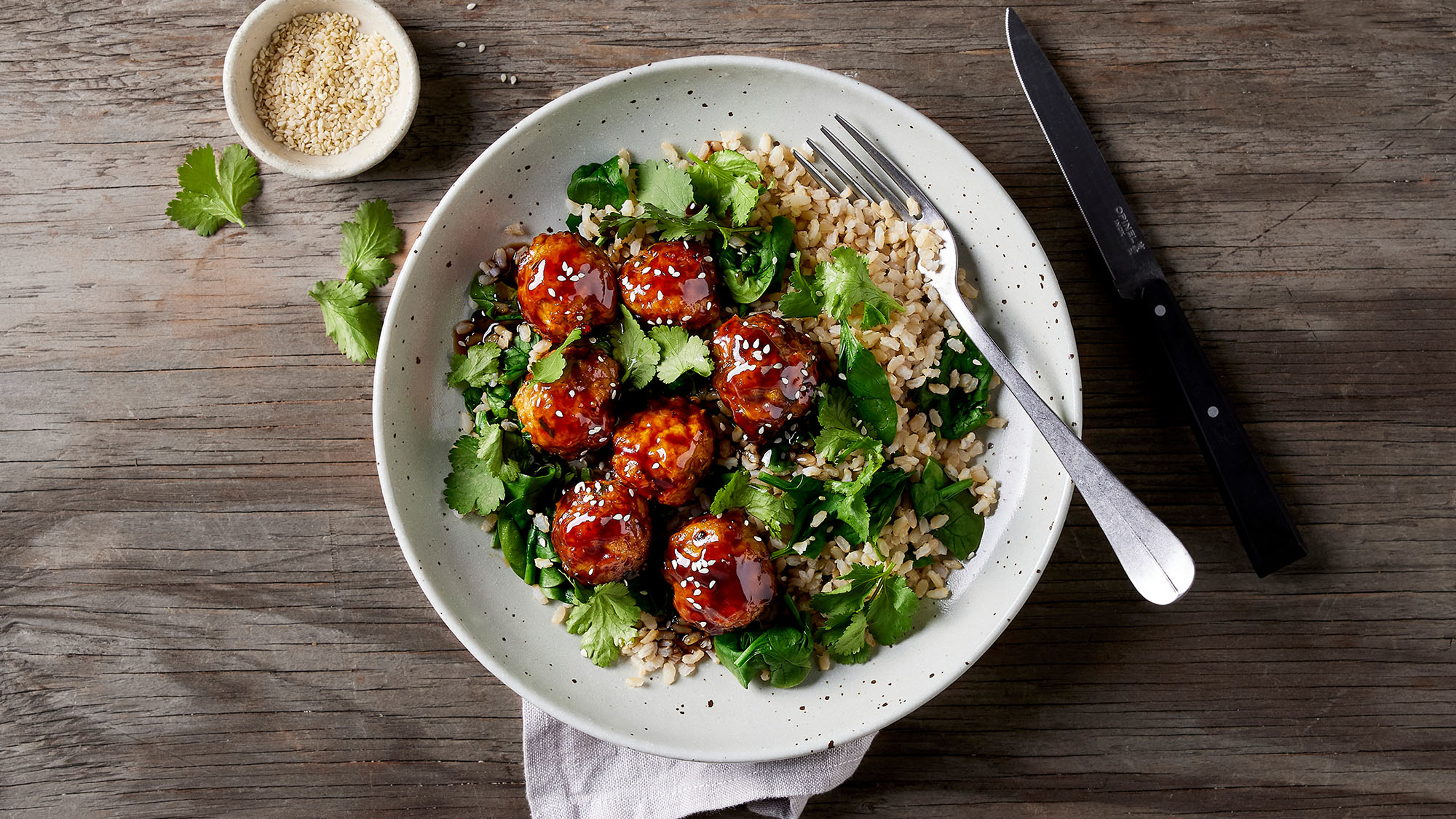 A bowl of sticky blazed chicken meatballs, sprinkled with cilantro leaves, sits on a wooden table, ready for eating.