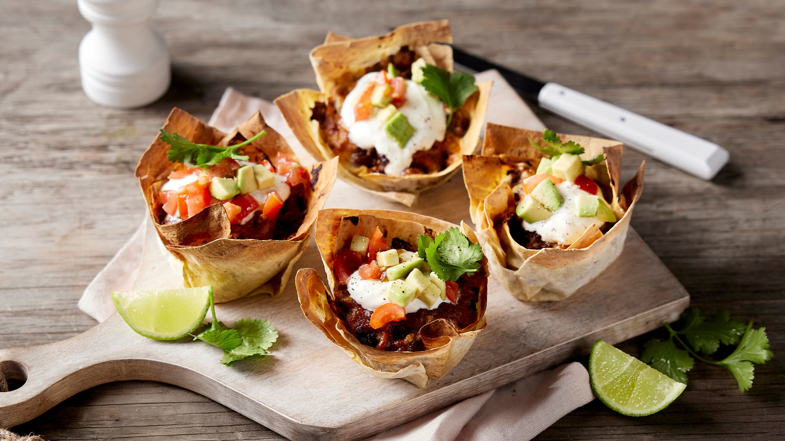 A serve of 4 taco cups sit on a wooden cutting board, surrounded by lime quarters and cilantro leaves.
