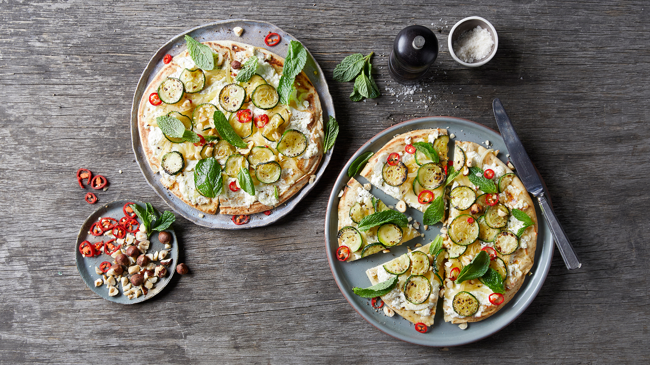 Two plates with Zucchini & Hazelnut Flatbreads on each sit on a wooden table, ready for eating.