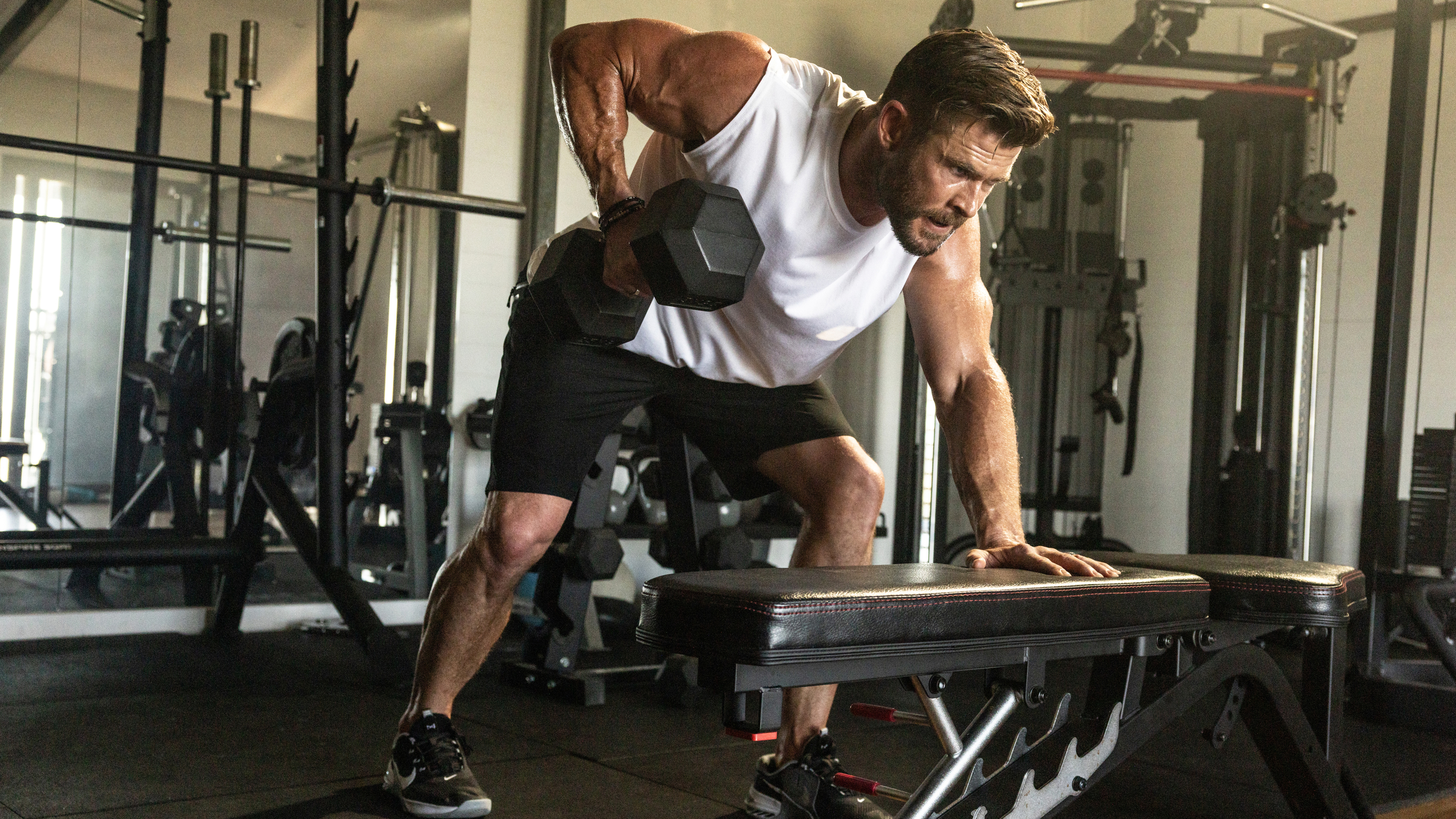 Chris Hemsworth performs bent over rows on the bench in a gym.
