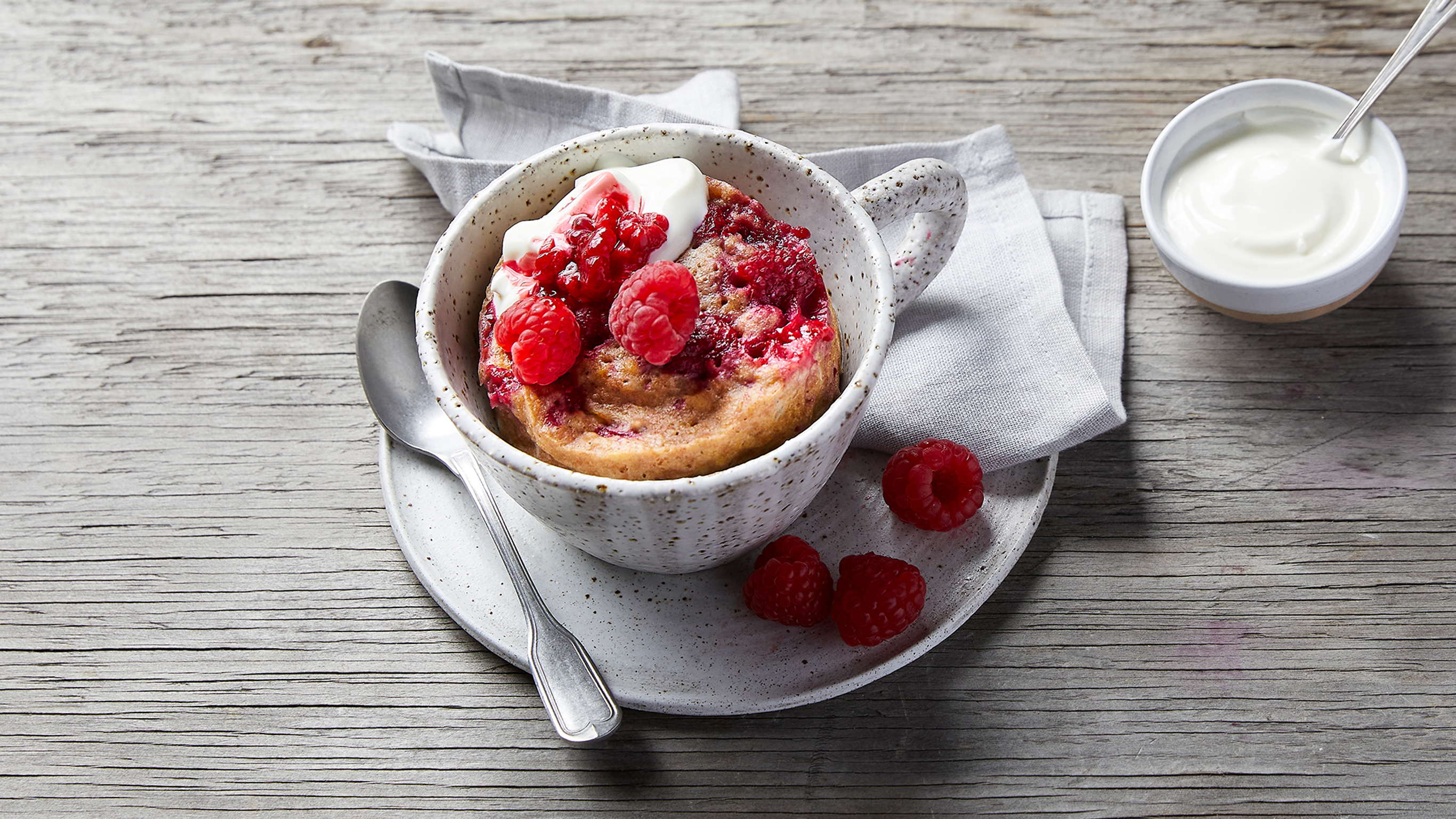 An overhead photo of Lemon & Raspberry Microwave Mug Cake in a speckled ceramic cup, served  with raspberries.